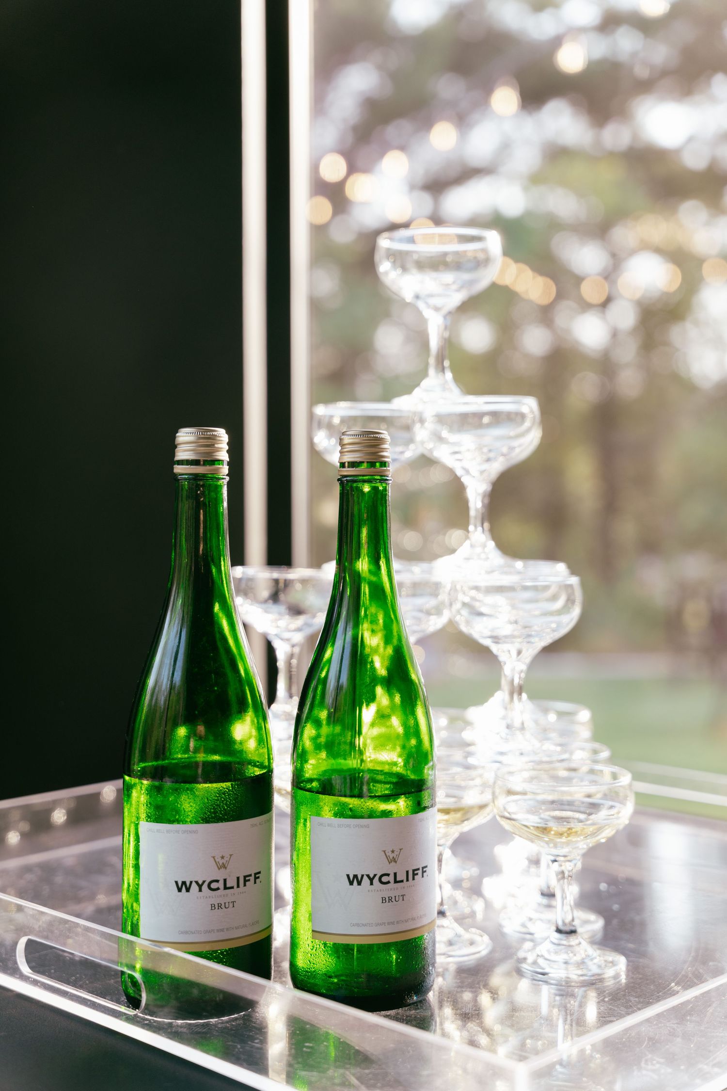 Two green Wycliff Brut champagne bottles beside stacked crystal coupe glasses on a silver tray at wedding.