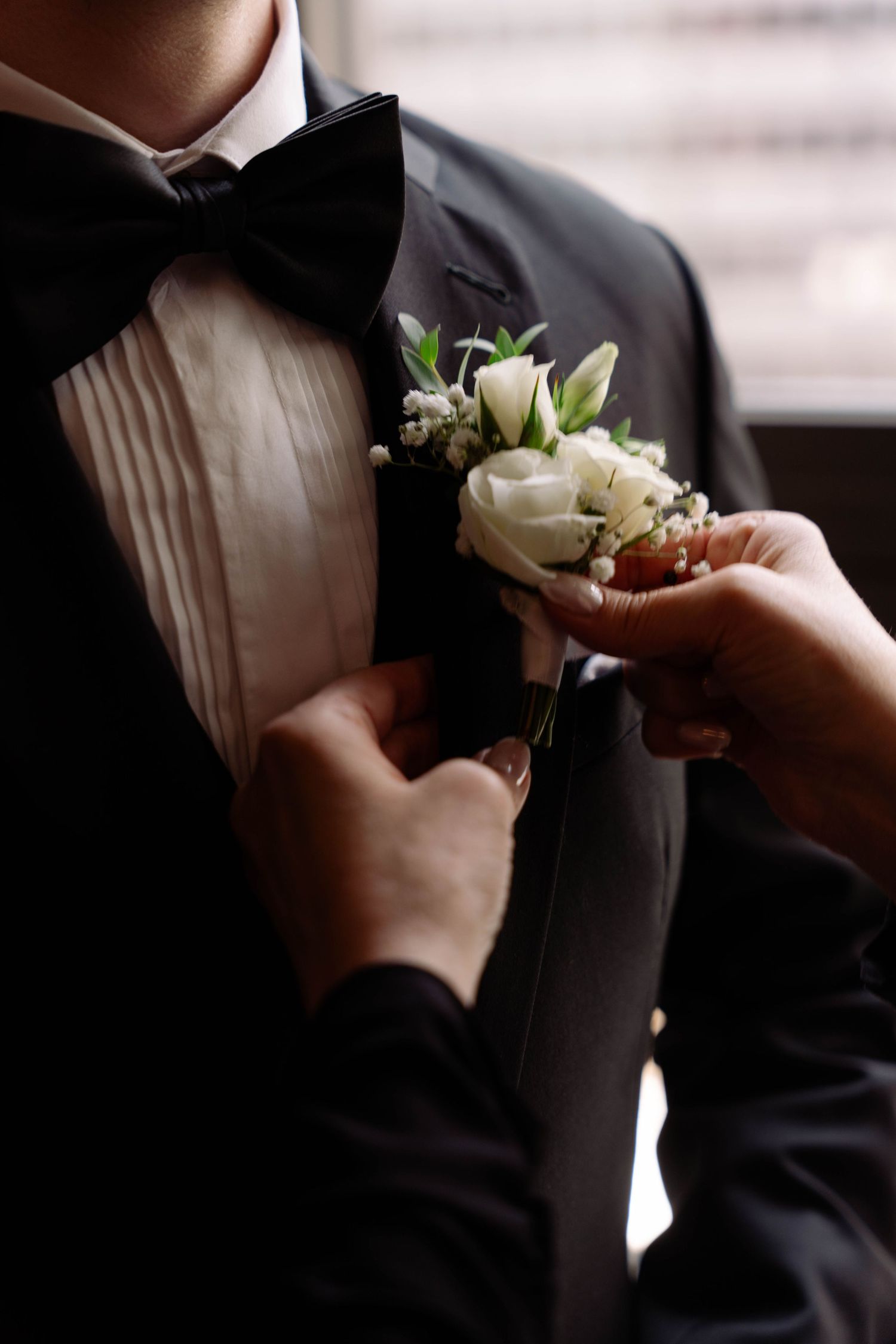 Hands pinning a white rose boutonniere onto a groom's black tuxedo lapel with bow tie.