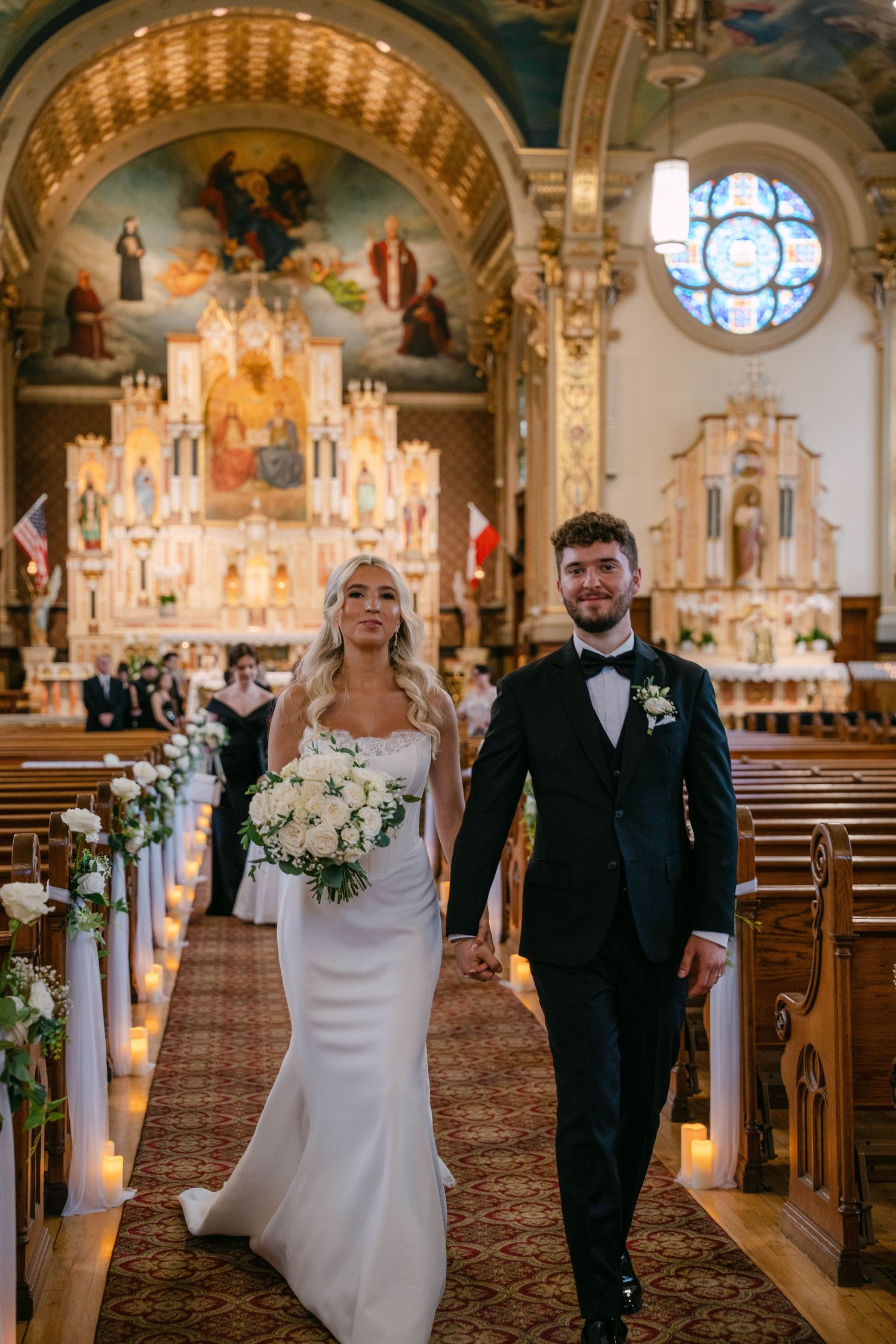 Smiling bride and groom walking hand in hand down a candlelit church aisle after their ceremony.