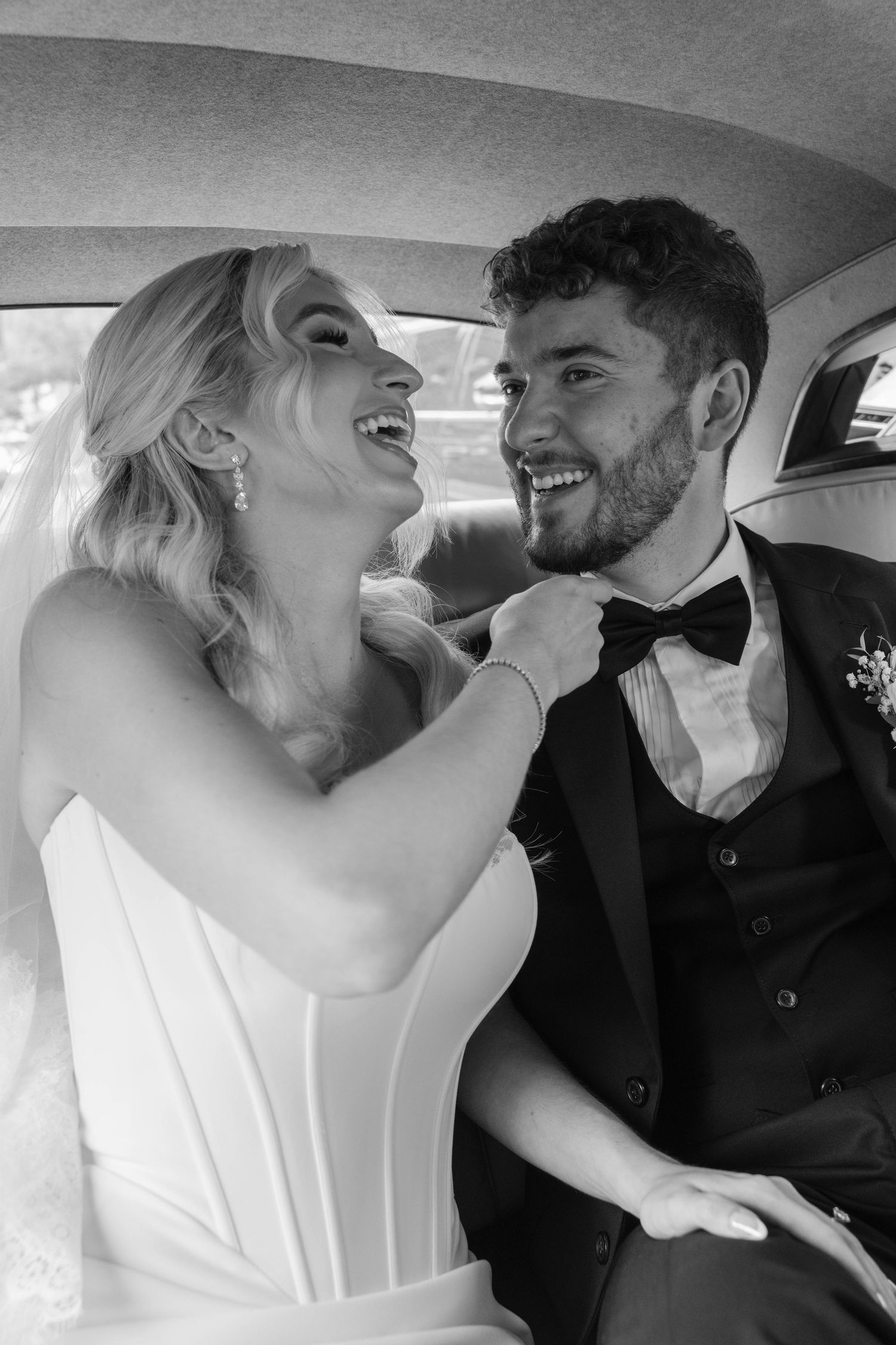 Bride and groom laughing together in car backseat, bride adjusting grooms bow tie, black and white.