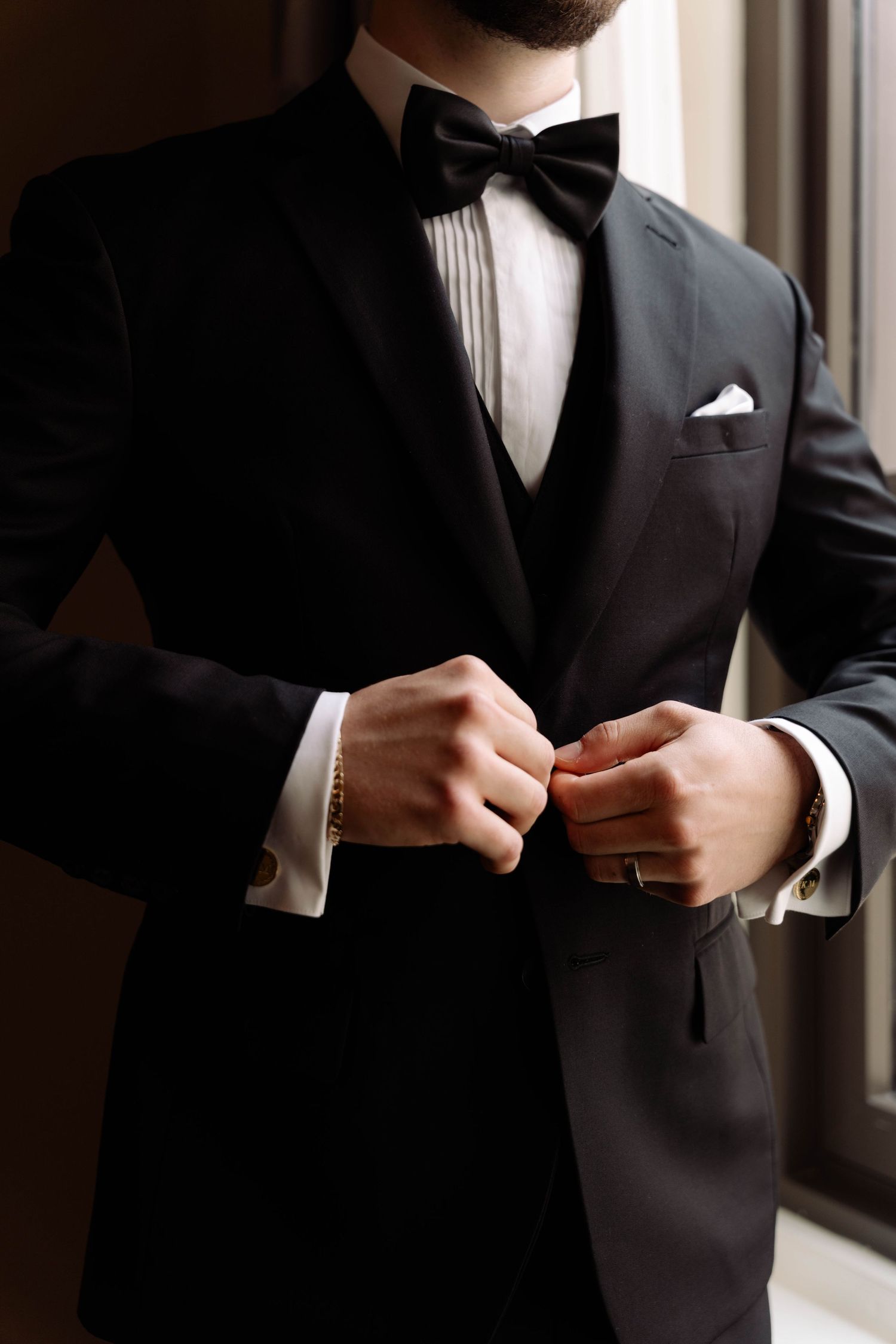 Groom in black tuxedo with bow tie buttoning jacket near window.