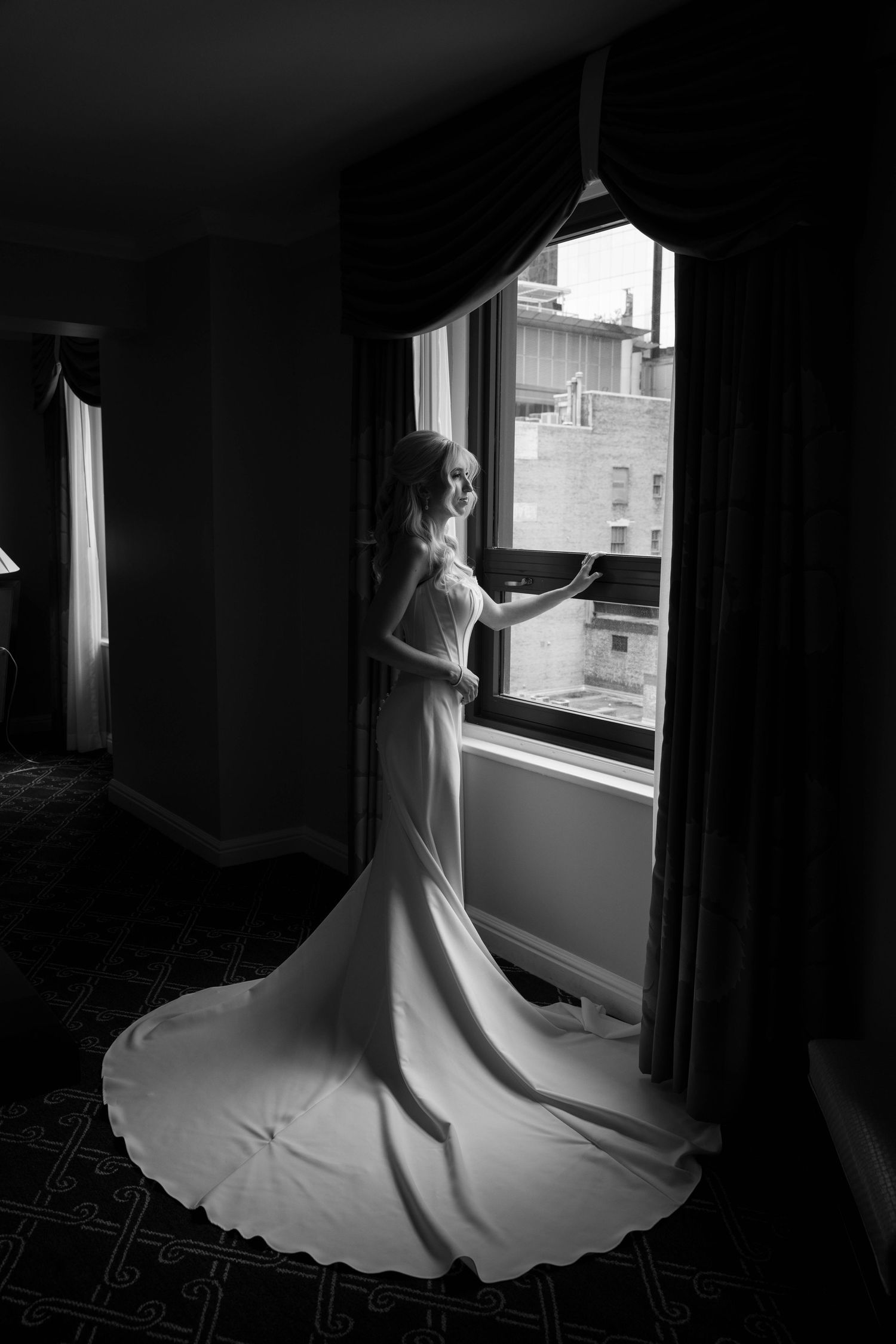 Bride in flowing white gown stands by hotel window in dramatic black and white wedding portrait.