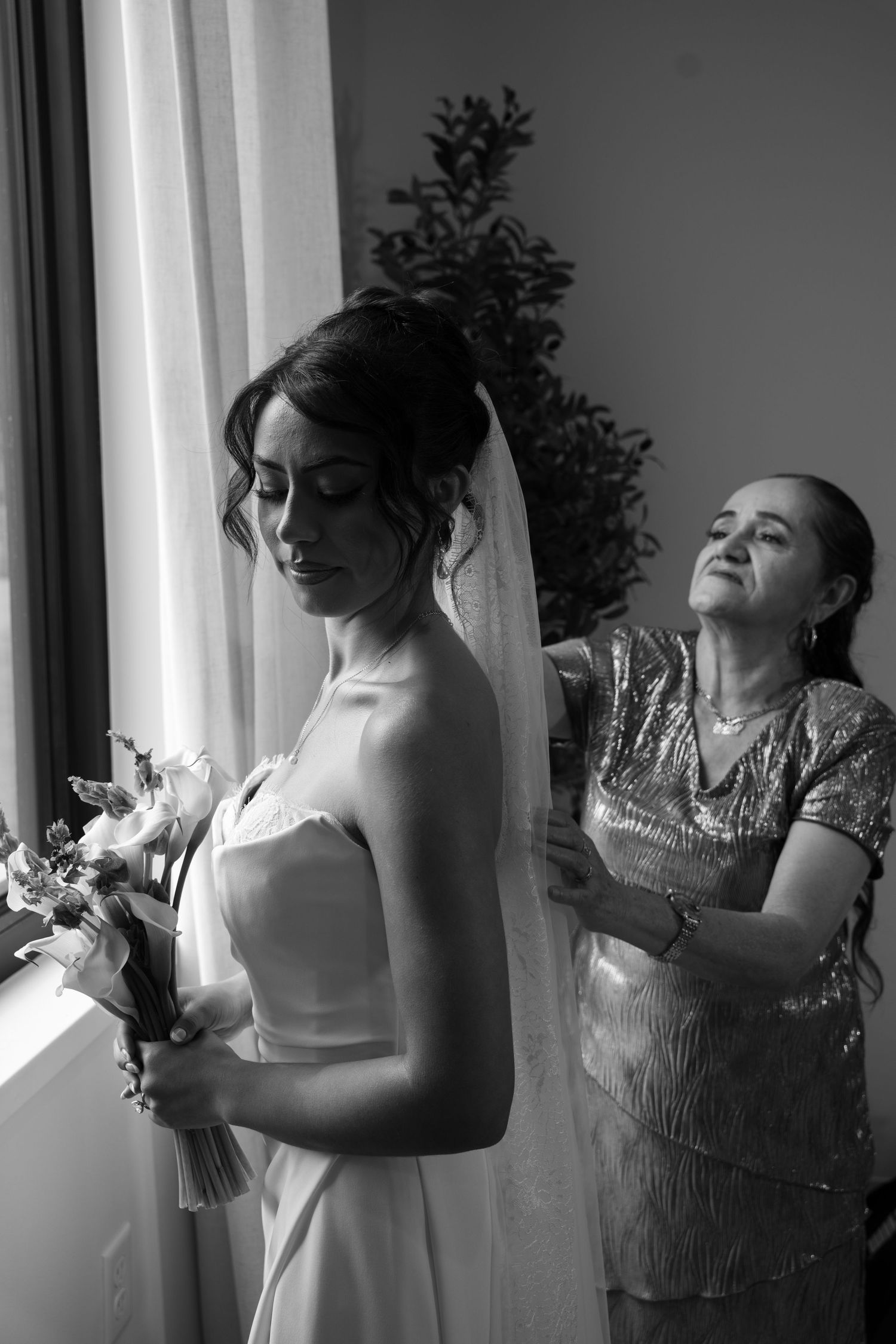 Black and white photo of mother helping bride with dress near window before wedding.