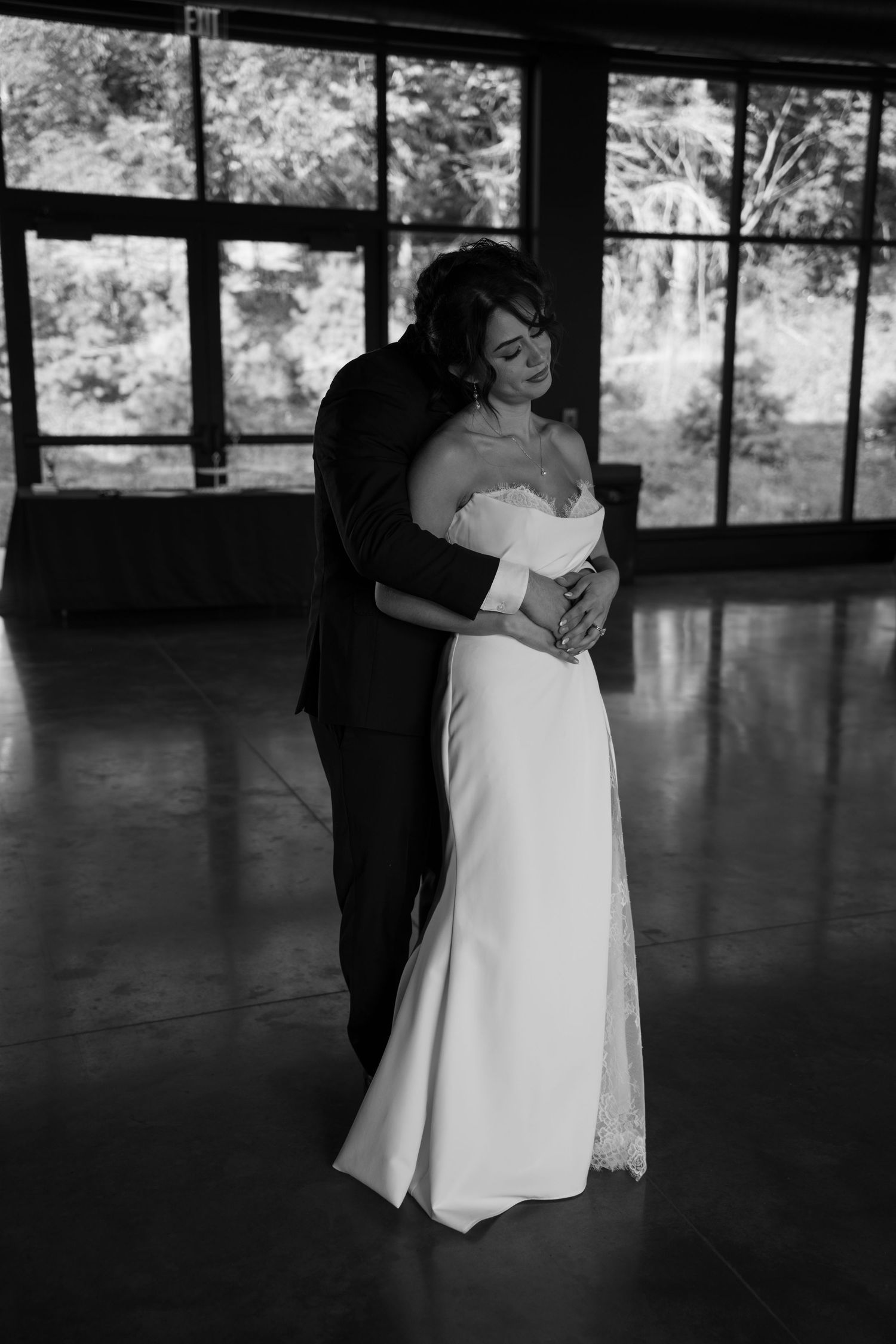 Bride and groom slow dance together in elegant black and white wedding photo inside a glass-windowed venue.