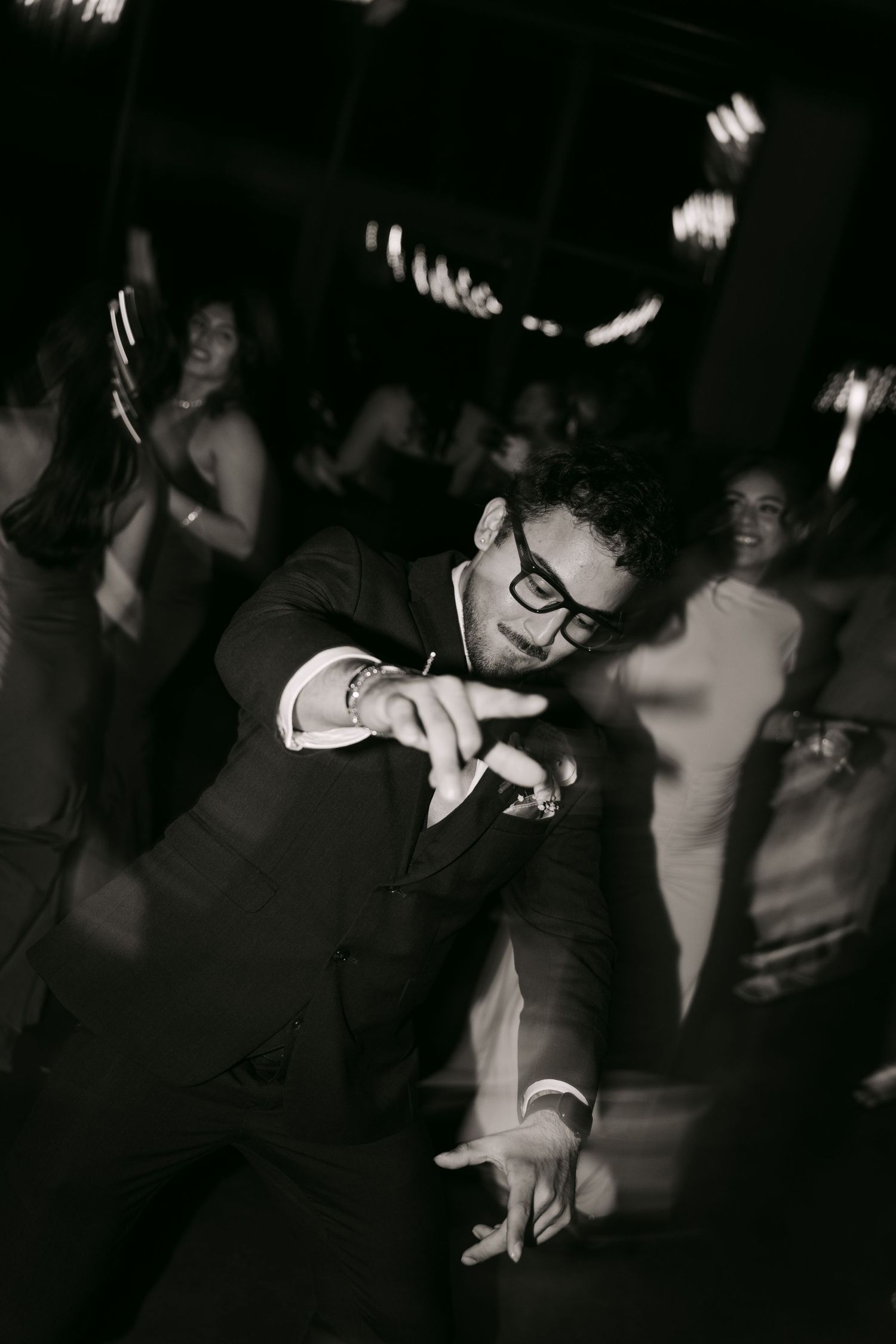 Black and white photo of groom dancing energetically at wedding reception with guests in background.