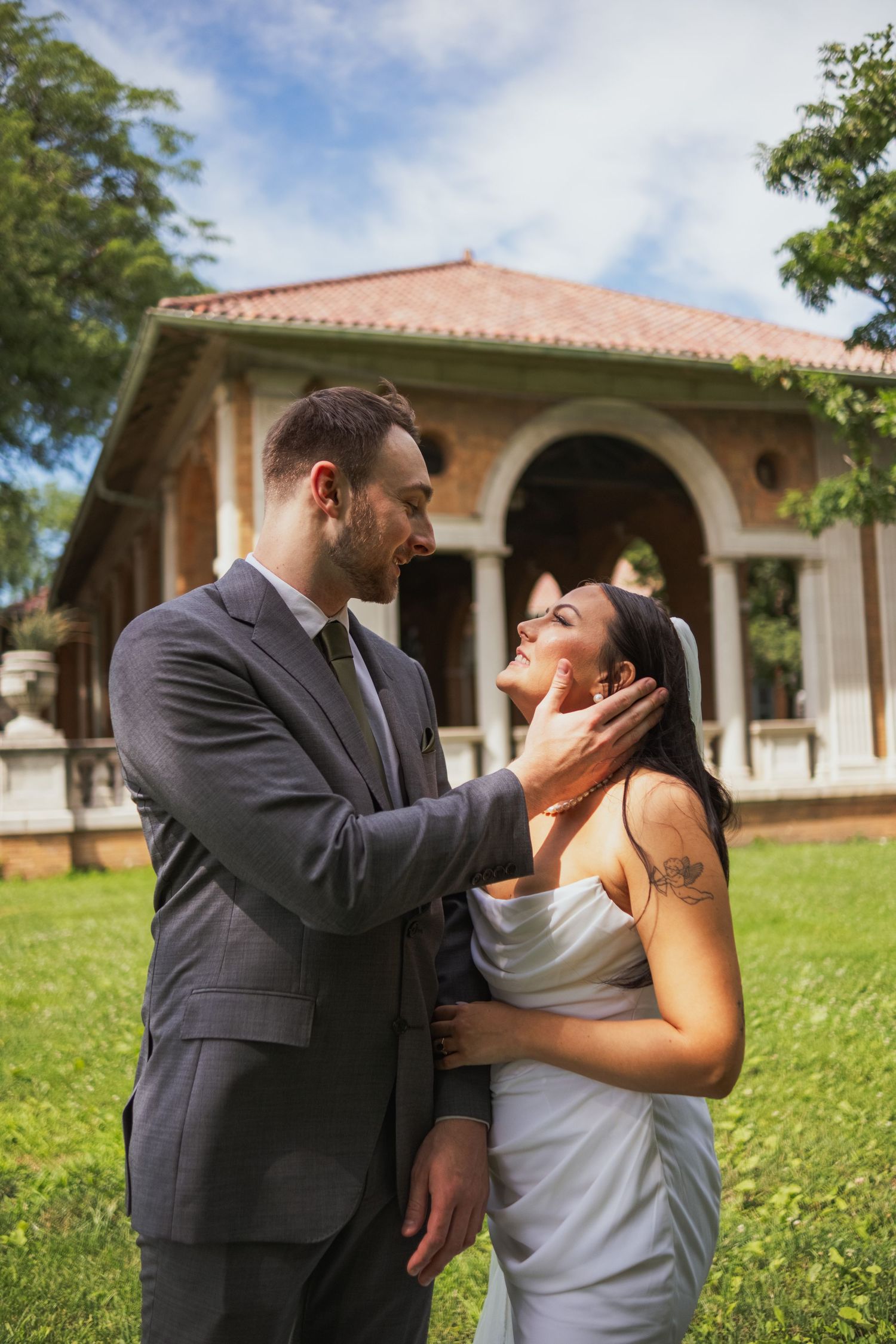 Groom in gray suit tenderly holding bride's face outdoors near historic building.