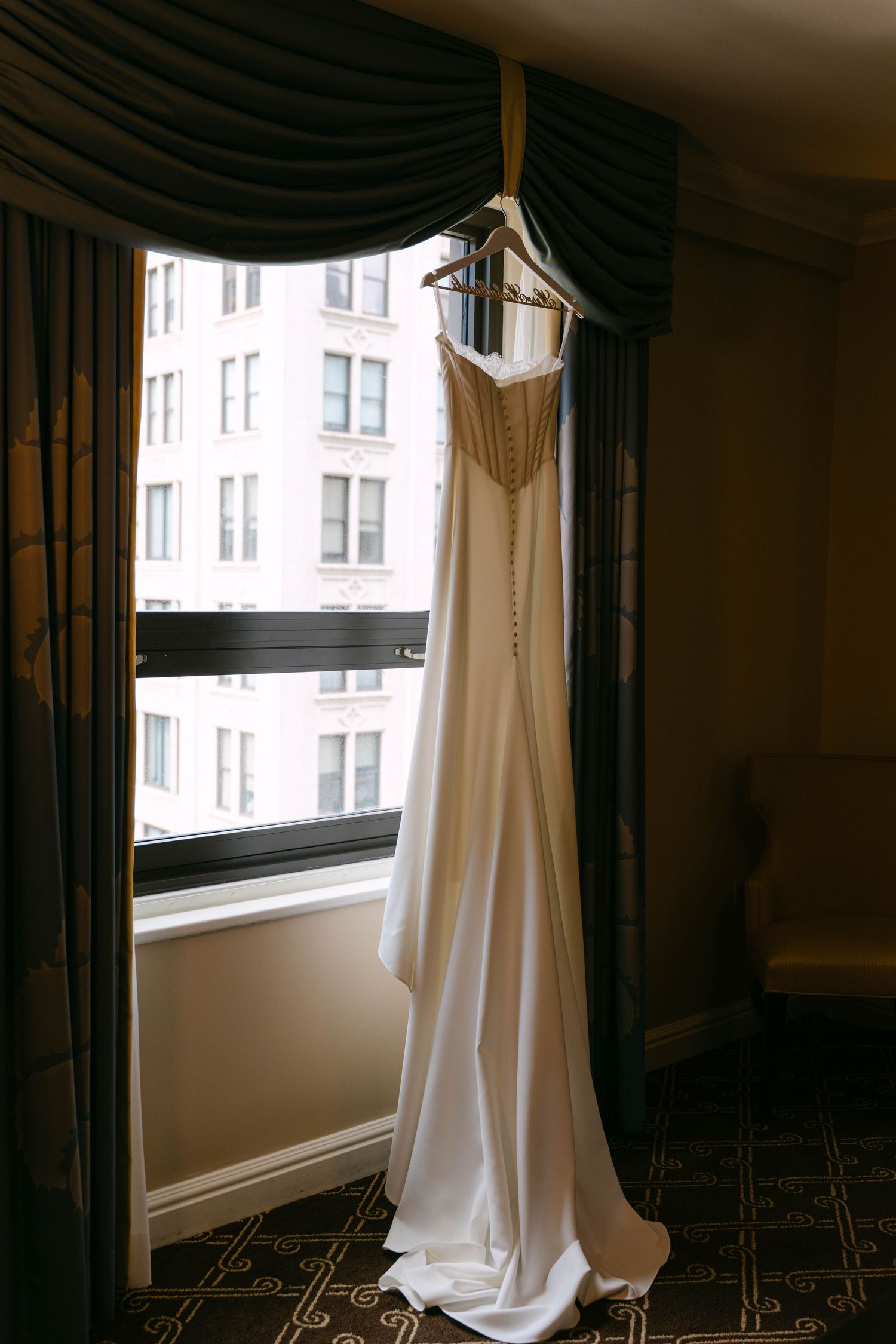 Elegant white wedding gown hanging on a hanger by a window in a hotel room with dark curtains.