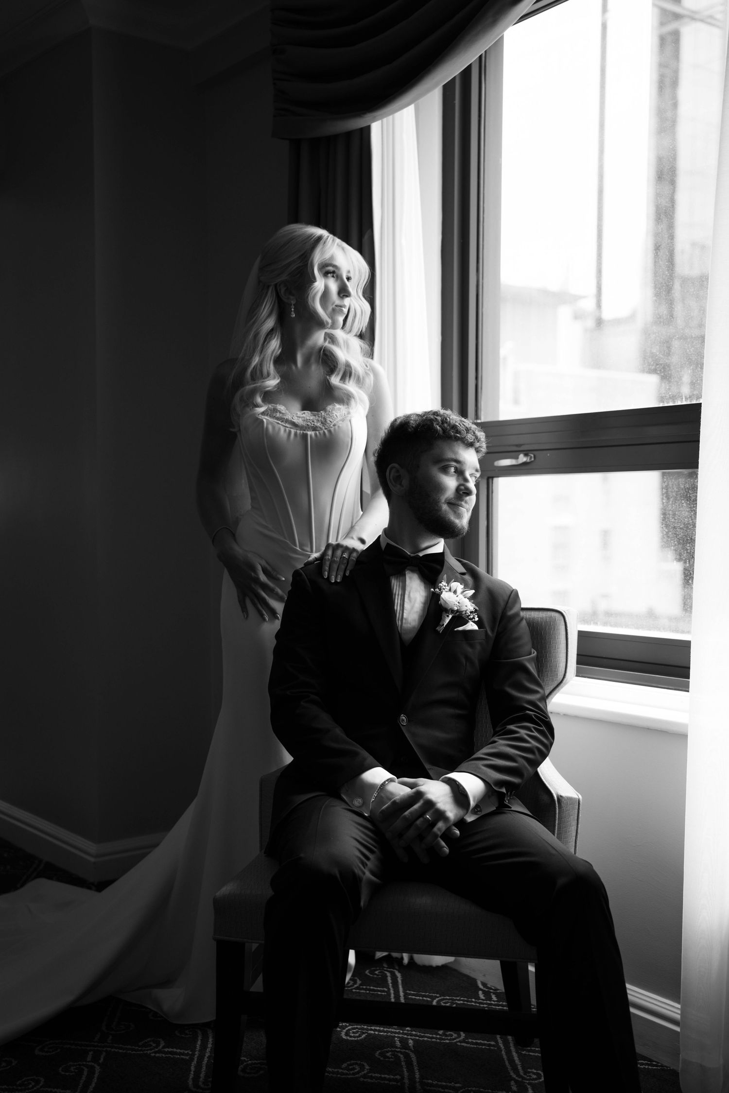 Bride and groom posing by hotel window in black and white, groom seated in chair, bride standing behind.