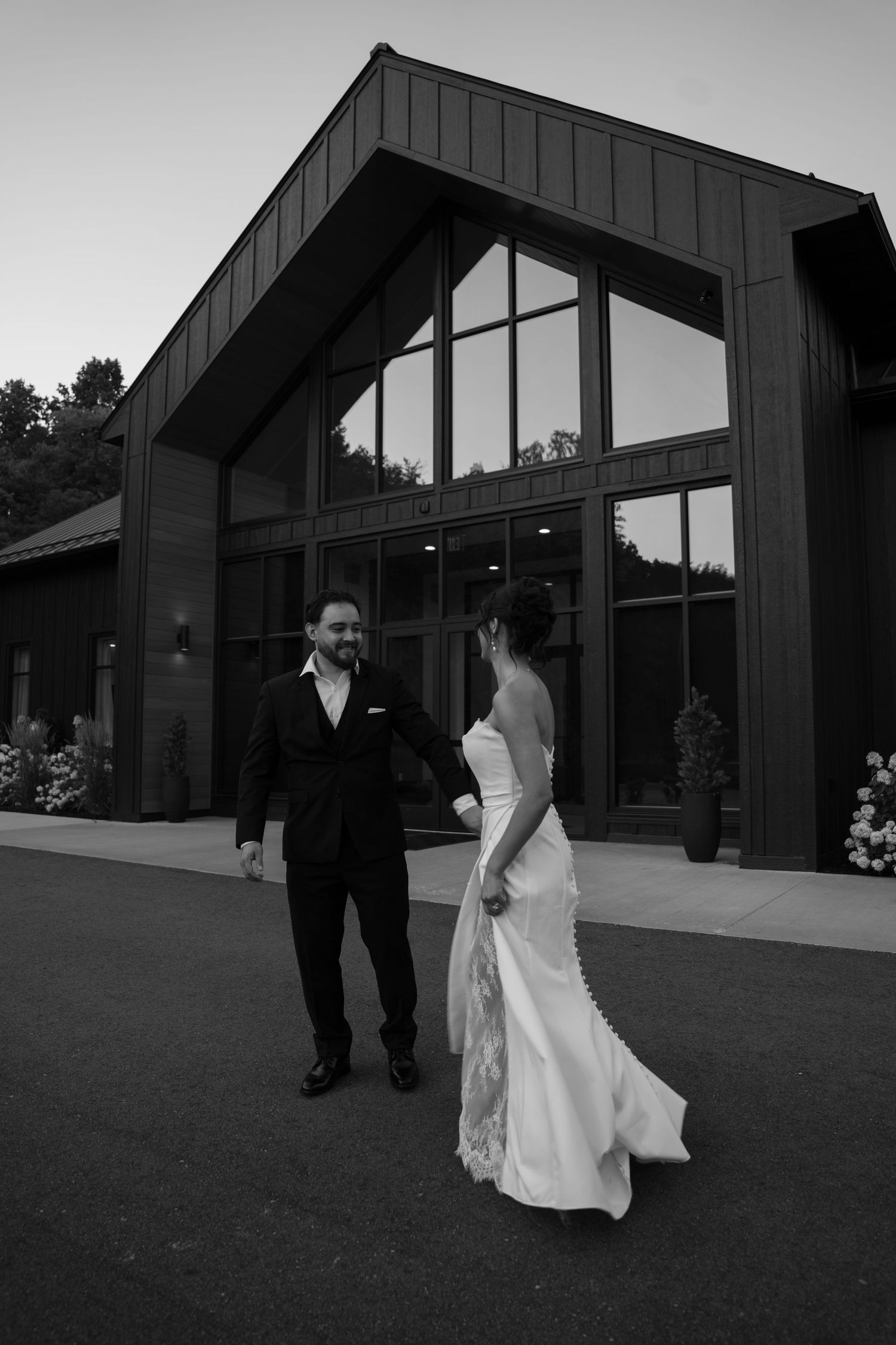 Bride and groom walk hand in hand outside an elegant modern venue in a black and white wedding photo.