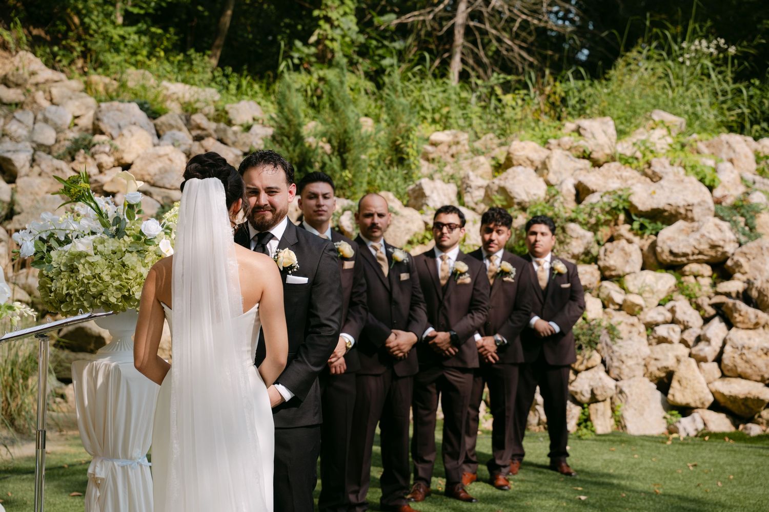 Groom faces bride at altar while groomsmen in brown suits stand in line at outdoor ceremony.