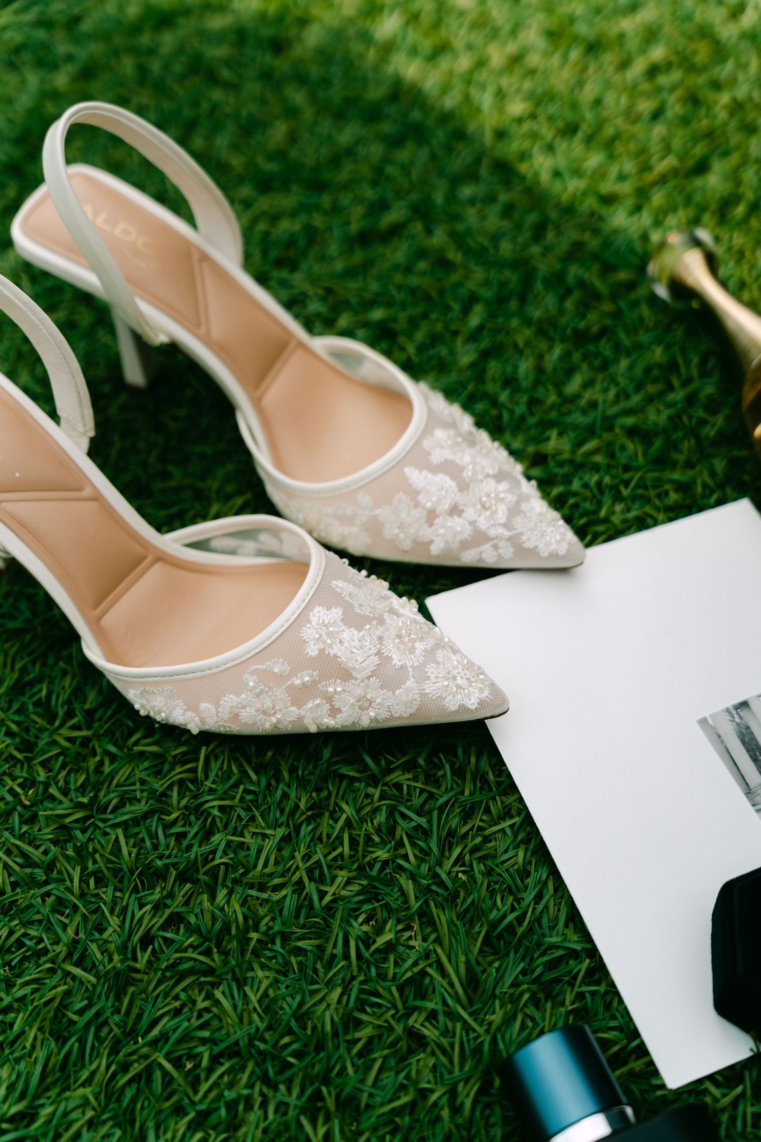 Elegant lace bridal slingback heels with floral applique resting on green grass beside wedding invitation.
