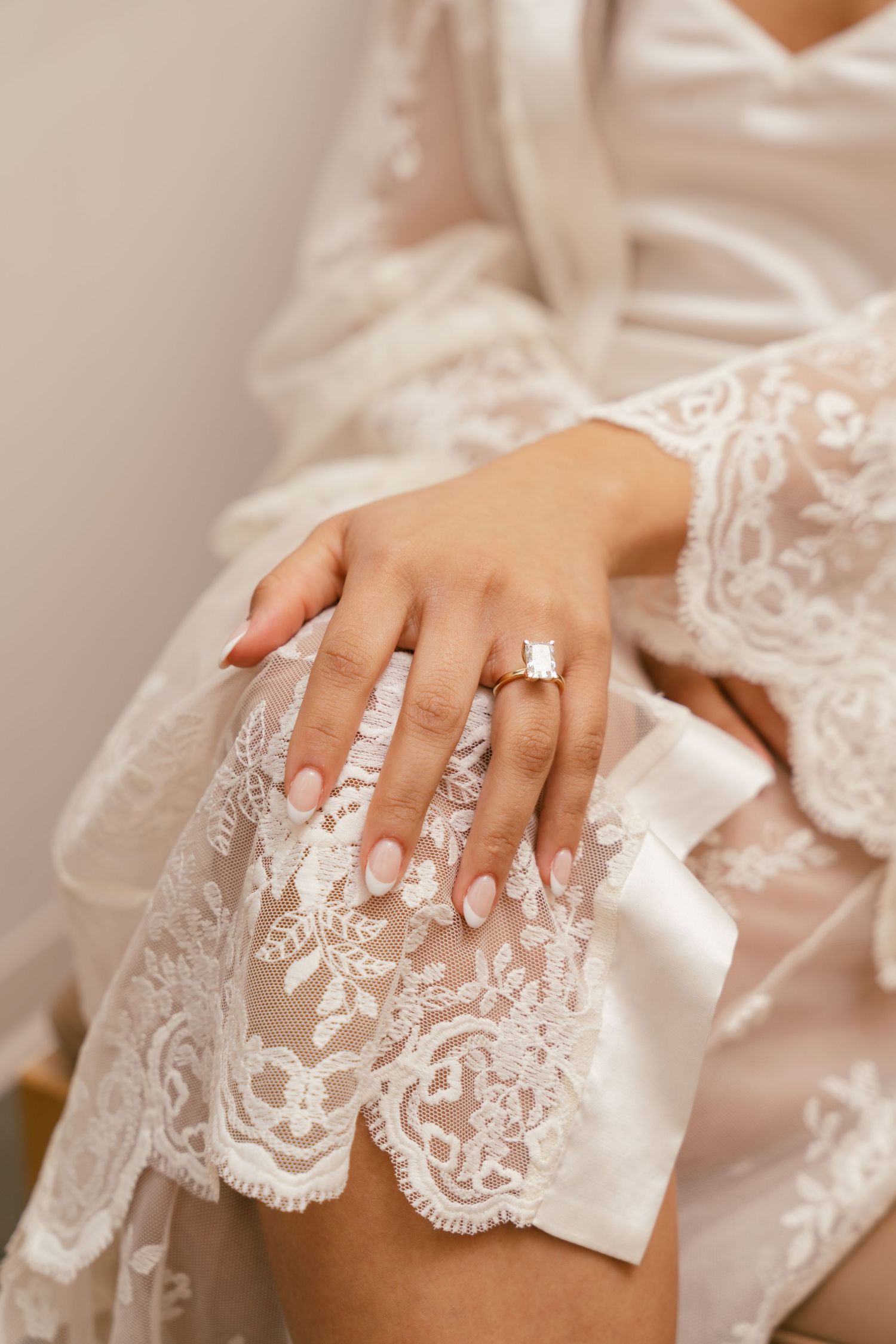 Close-up of brides hand wearing elegant square diamond engagement ring on white lace wedding robe.