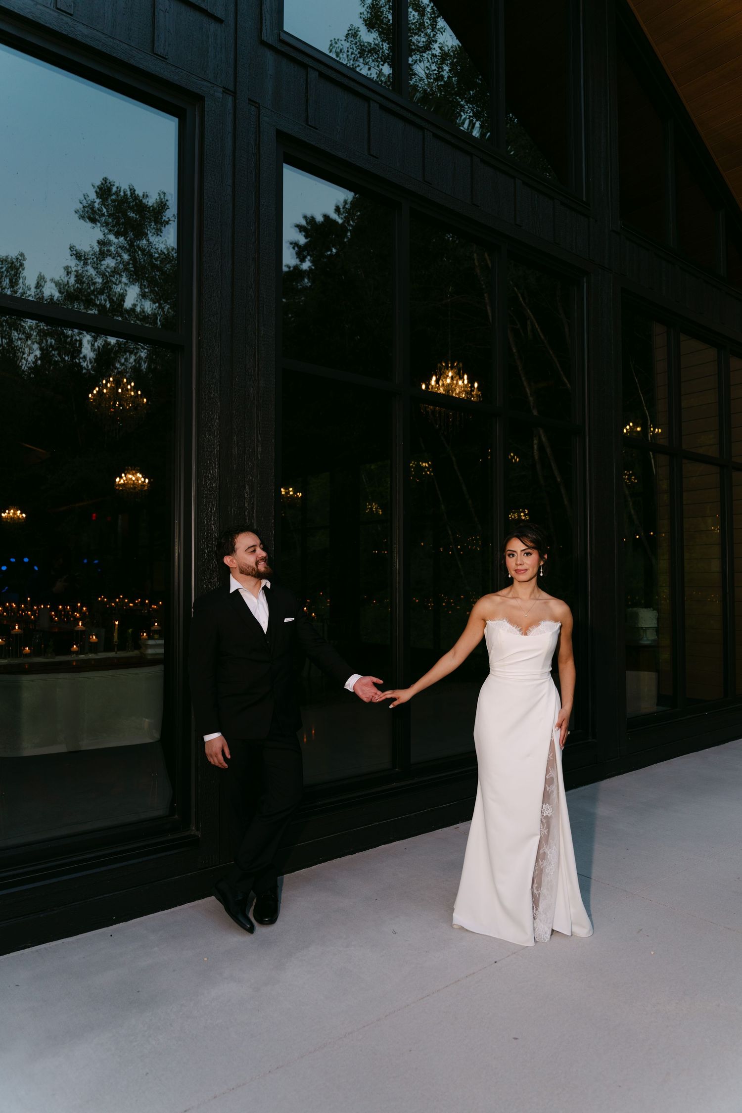 Bride in a sleek white lace gown holds grooms hand outside a stunning modern glass wedding venue.