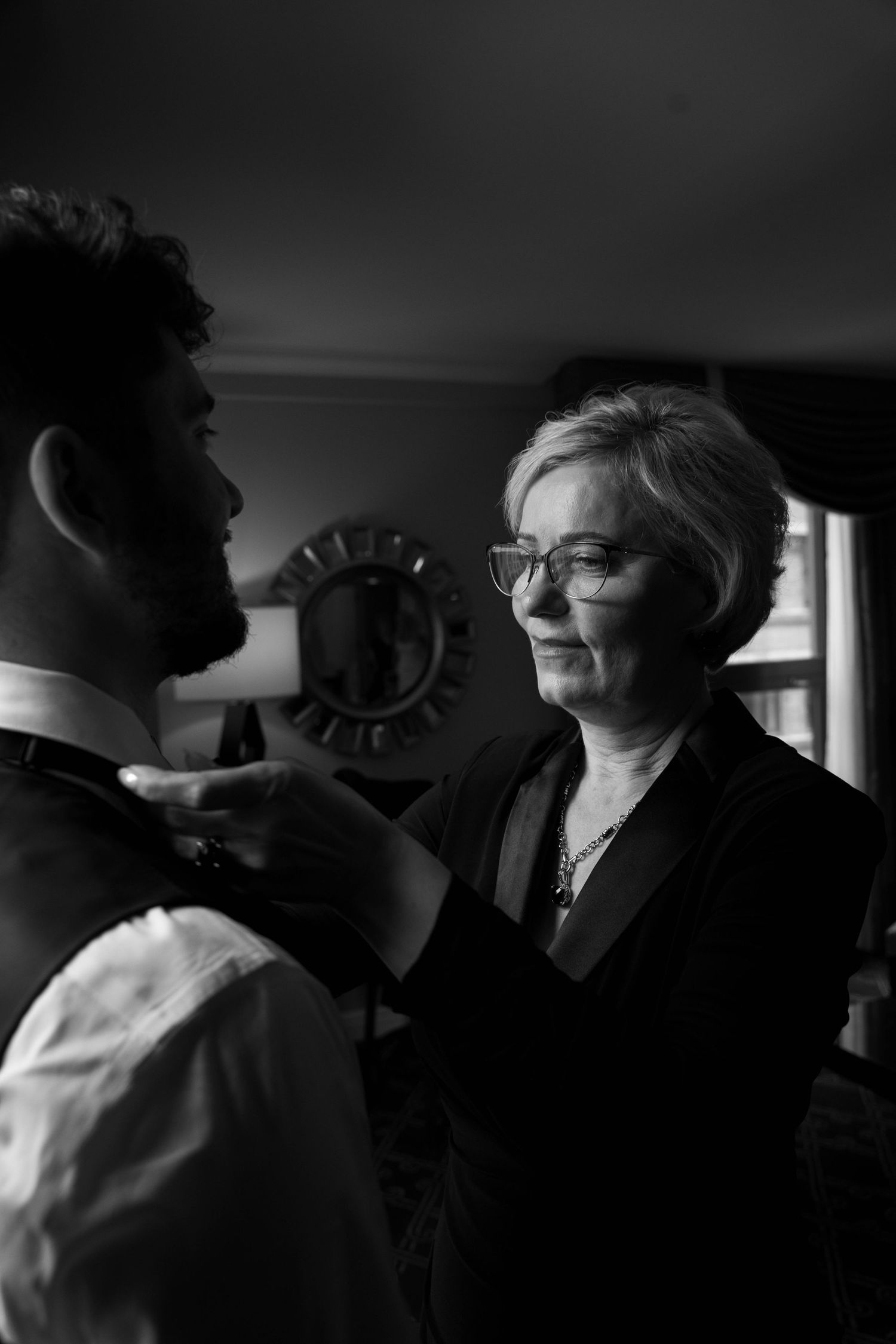 Black and white photo of a woman adjusting a mans bow tie, smiling warmly in an elegant indoor setting.