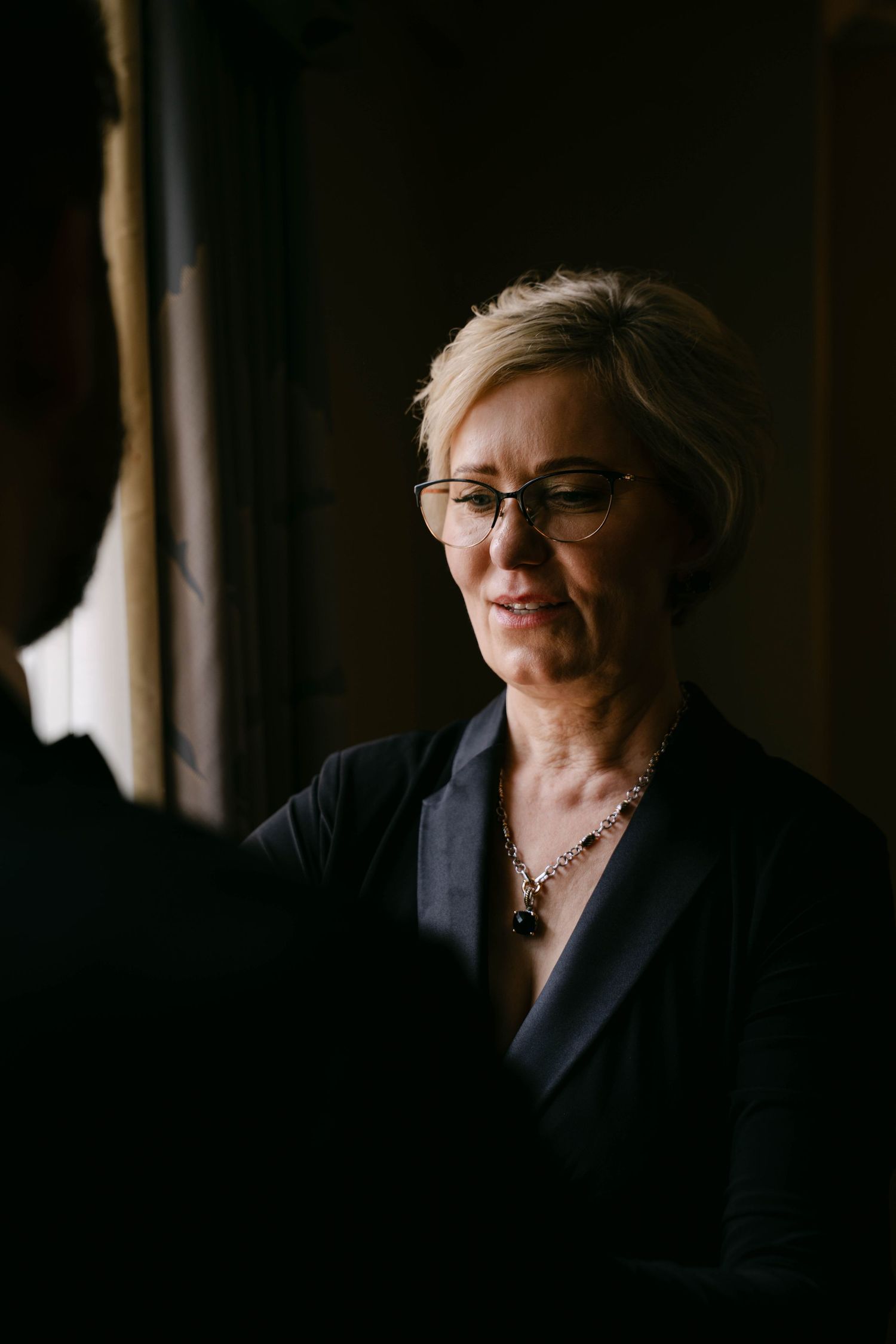 Elegant older woman with short blonde hair and glasses wearing a dark blouse and gemstone necklace at a wedding.
