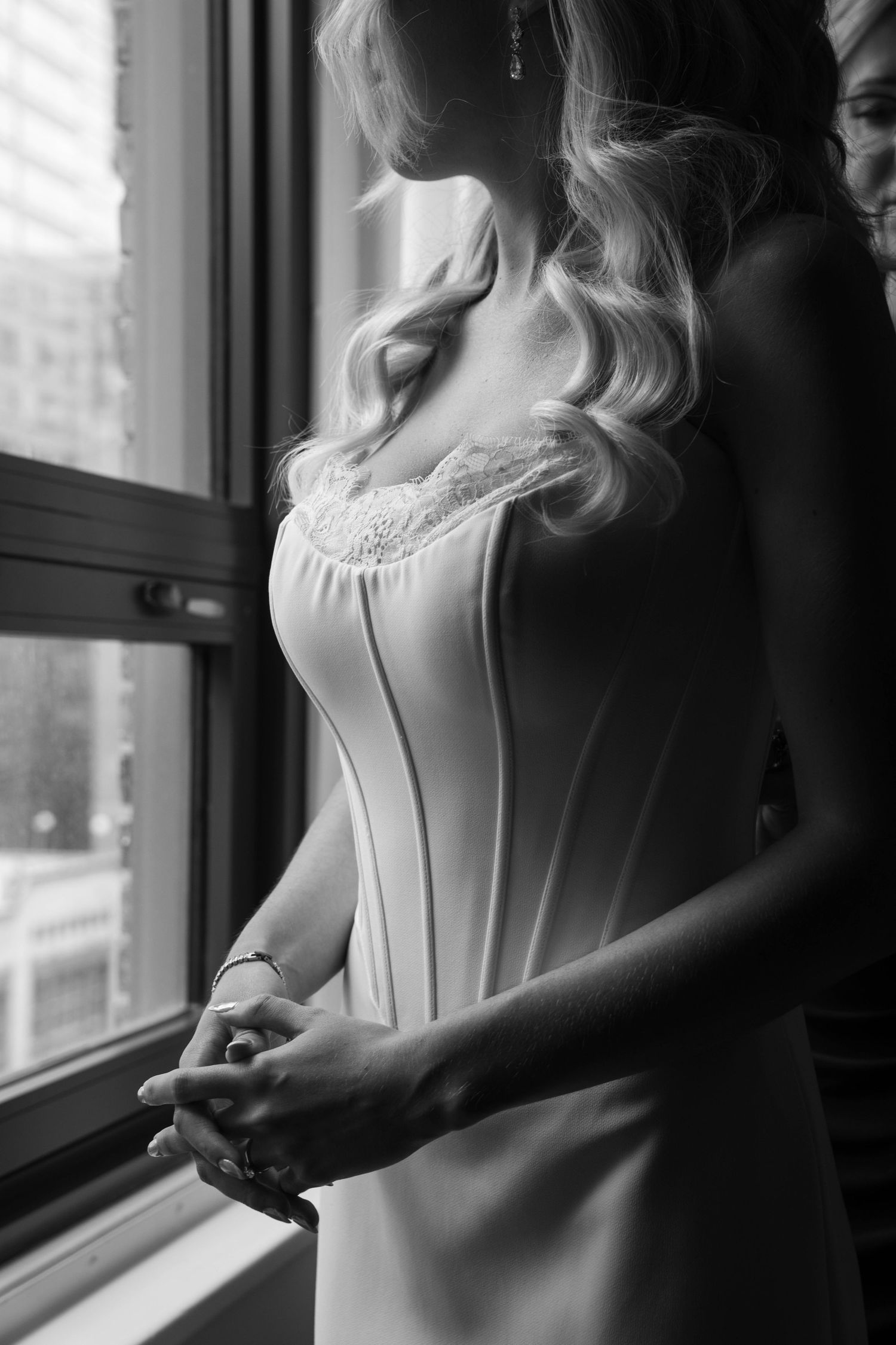 Elegant bride in white corset gown with lace detail standing by window in soft natural light.