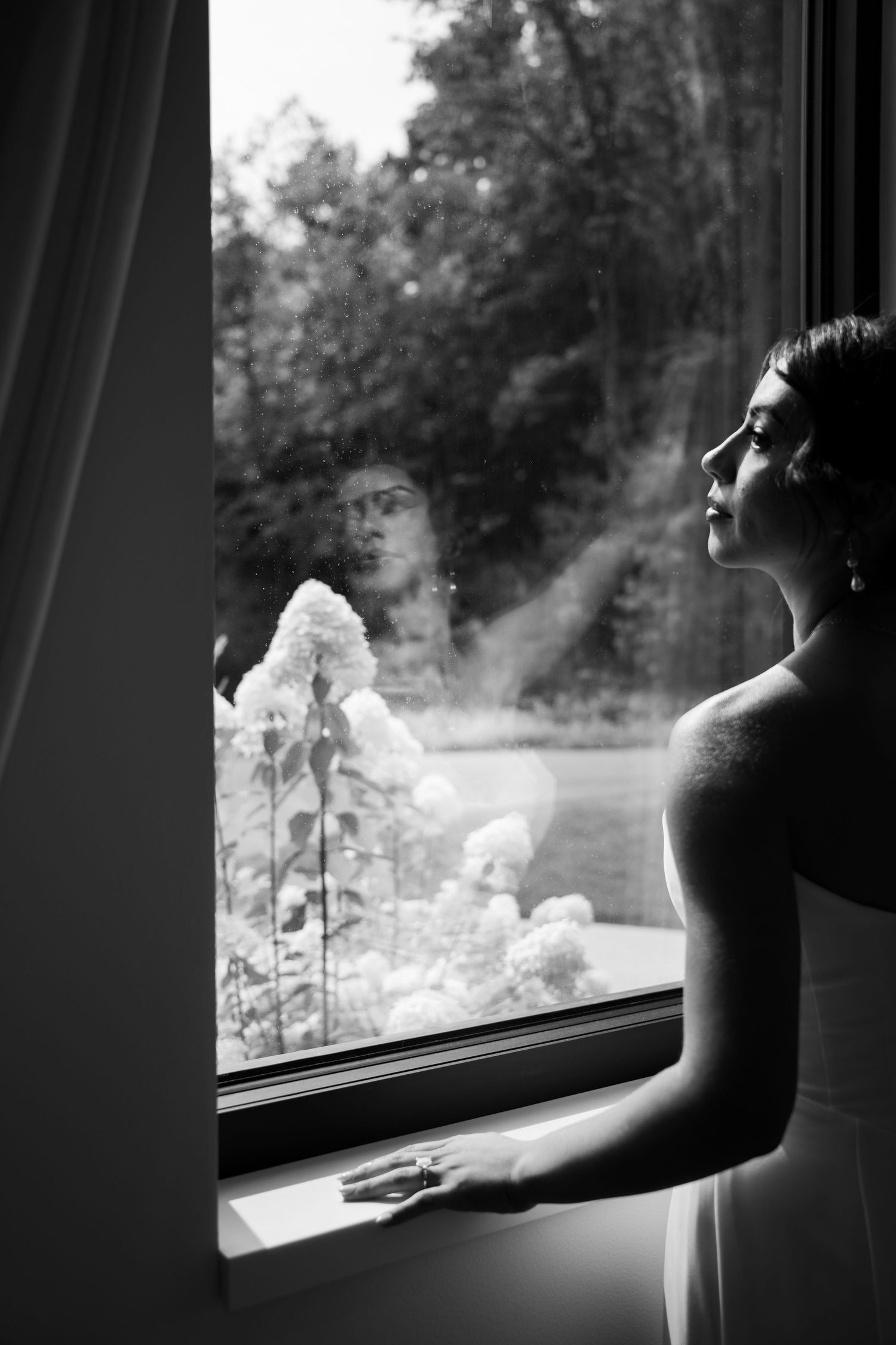 Black and white photo of bride gazing out window with reflection visible in glass.