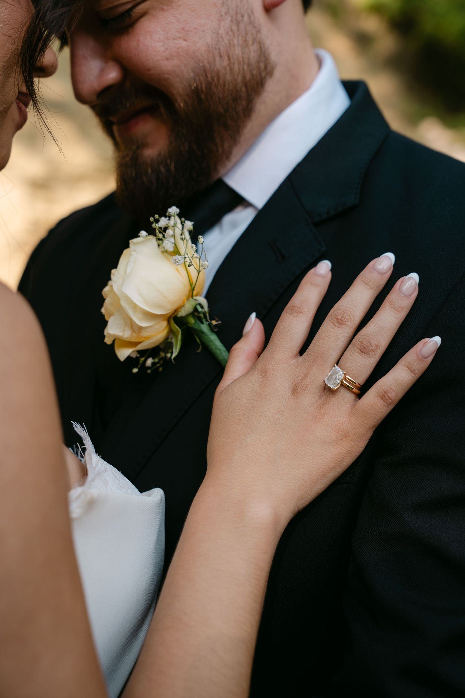 Close-up of bride's hand with oval diamond engagement ring resting on groom's black suit lapel.