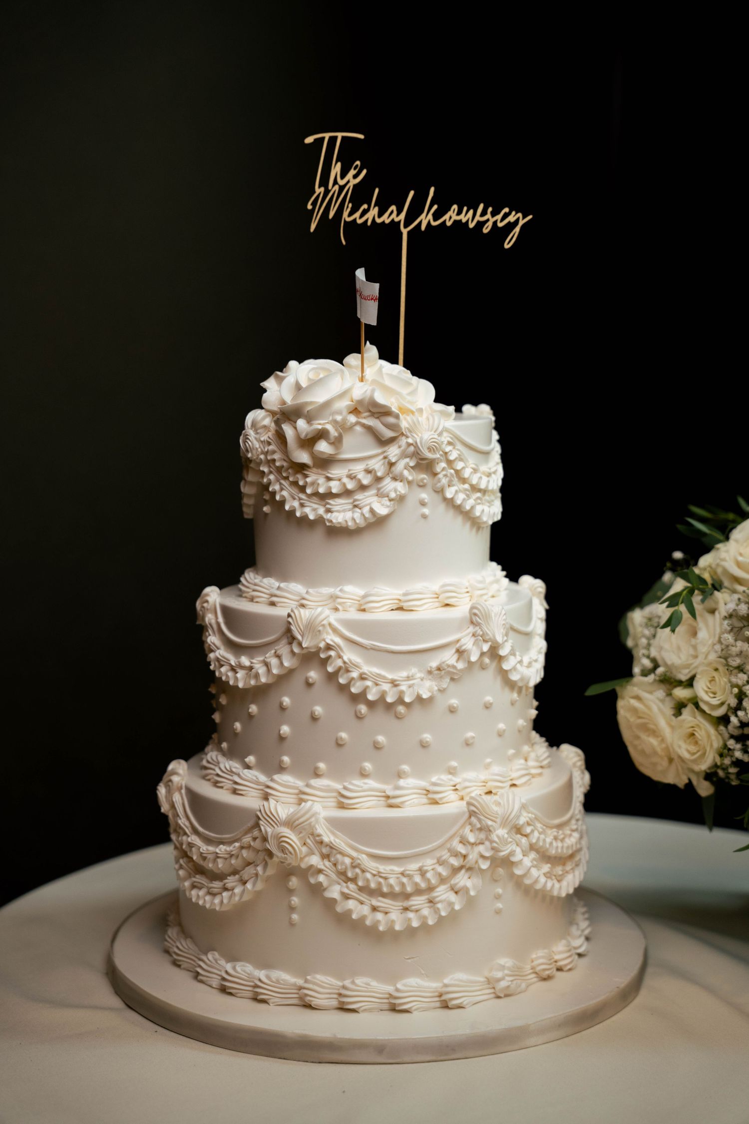 Elegant three-tier white wedding cake with pearl details and custom gold cake topper on dark background.