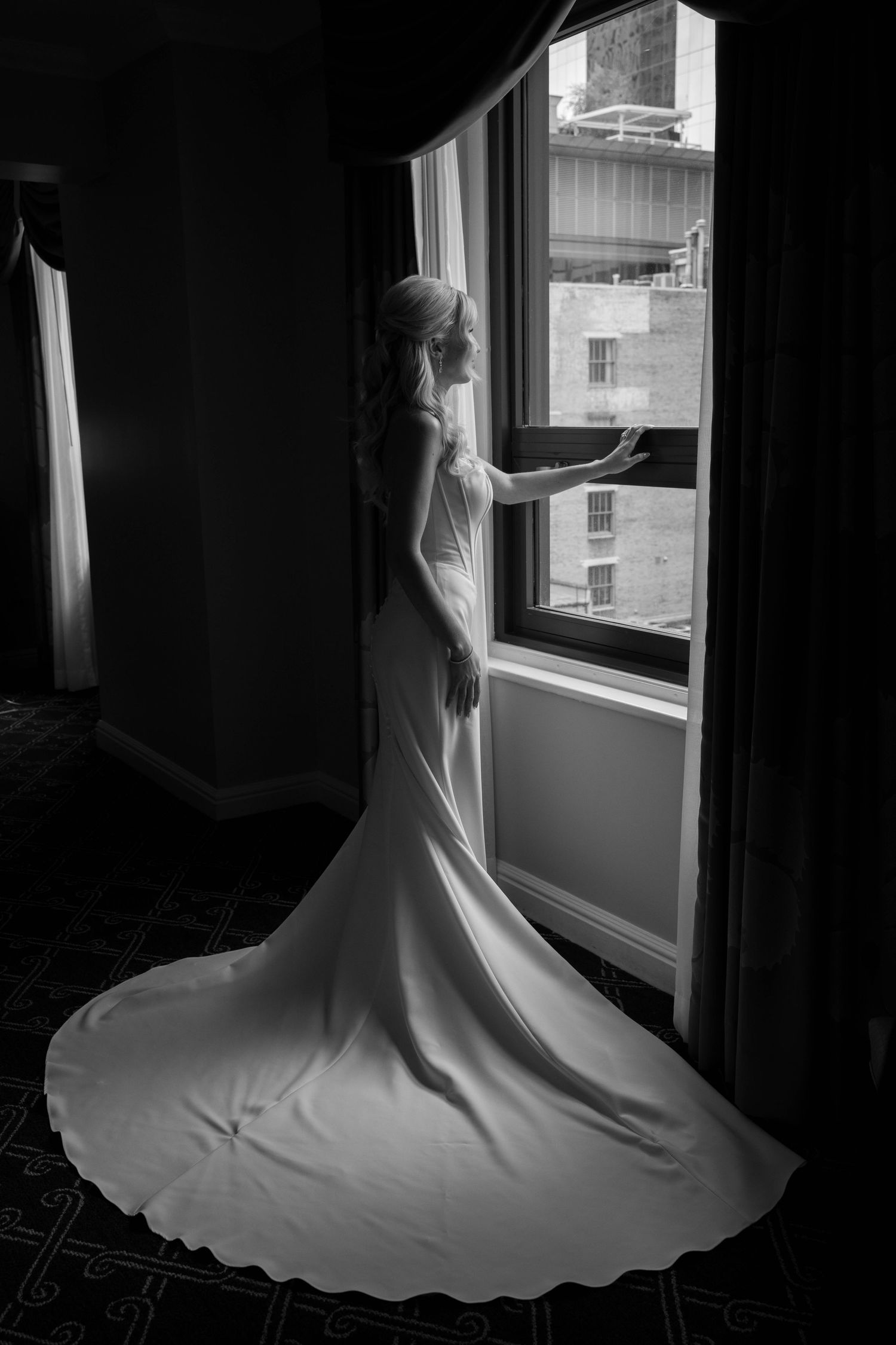 Bride in elegant white gown with long train stands by window in black and white wedding photo.