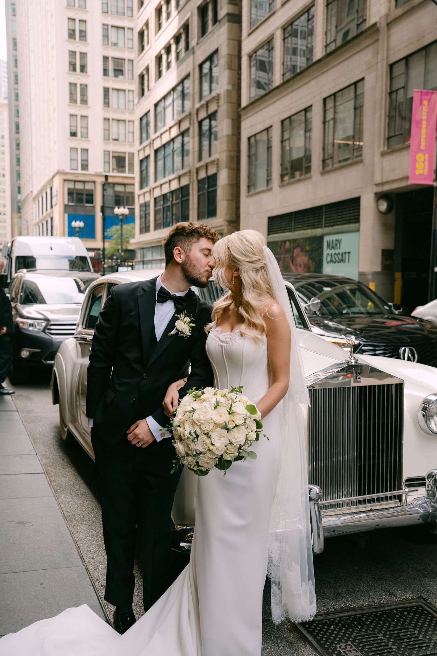 Bride and groom kissing beside vintage white Rolls Royce on city street.