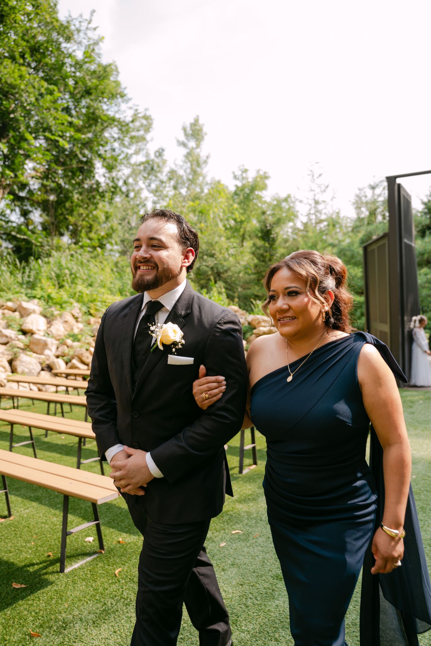 Groom and escort smile while walking down grass aisle at elegant outdoor wedding ceremony.