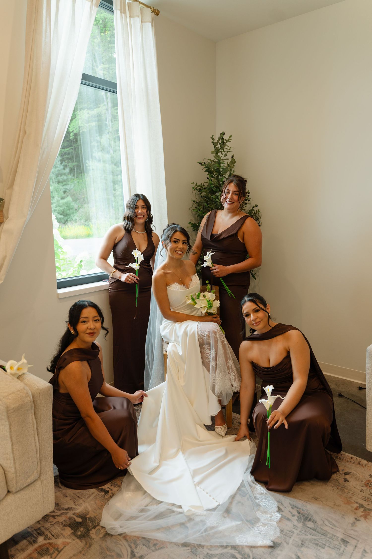 Bride in white gown surrounded by four bridesmaids in brown dresses holding calla lilies near window.