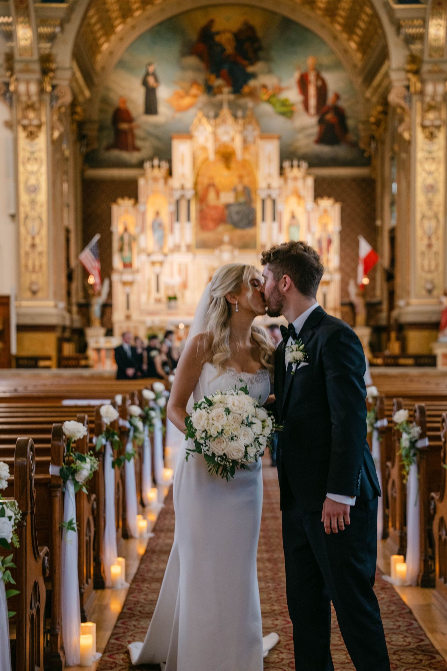 Bride and groom kissing at the altar of an ornate church with candlelit aisle and golden decor.