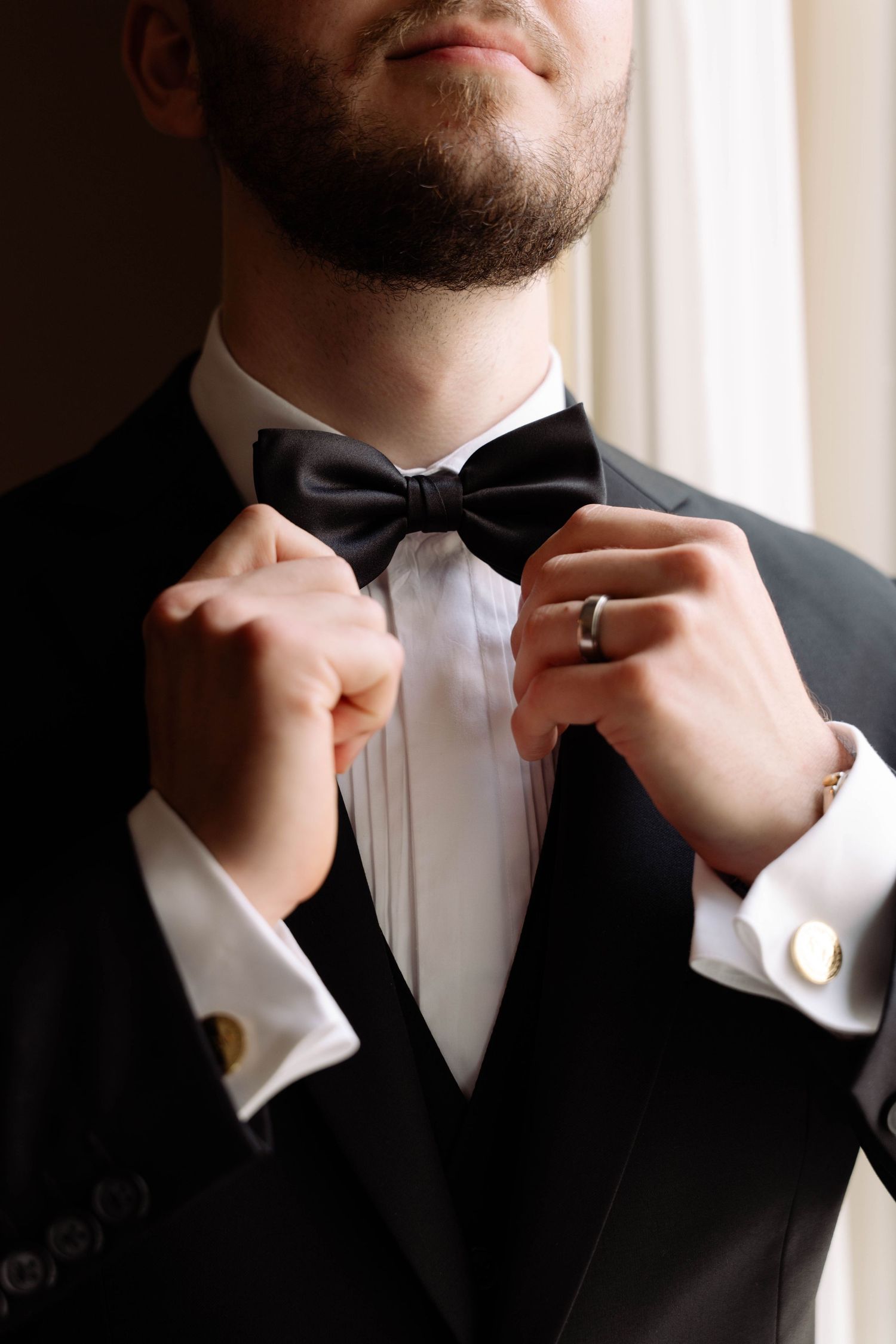 Groom in black tuxedo adjusting bow tie, wearing cufflinks and wedding ring, elegant formal attire for wedding day.