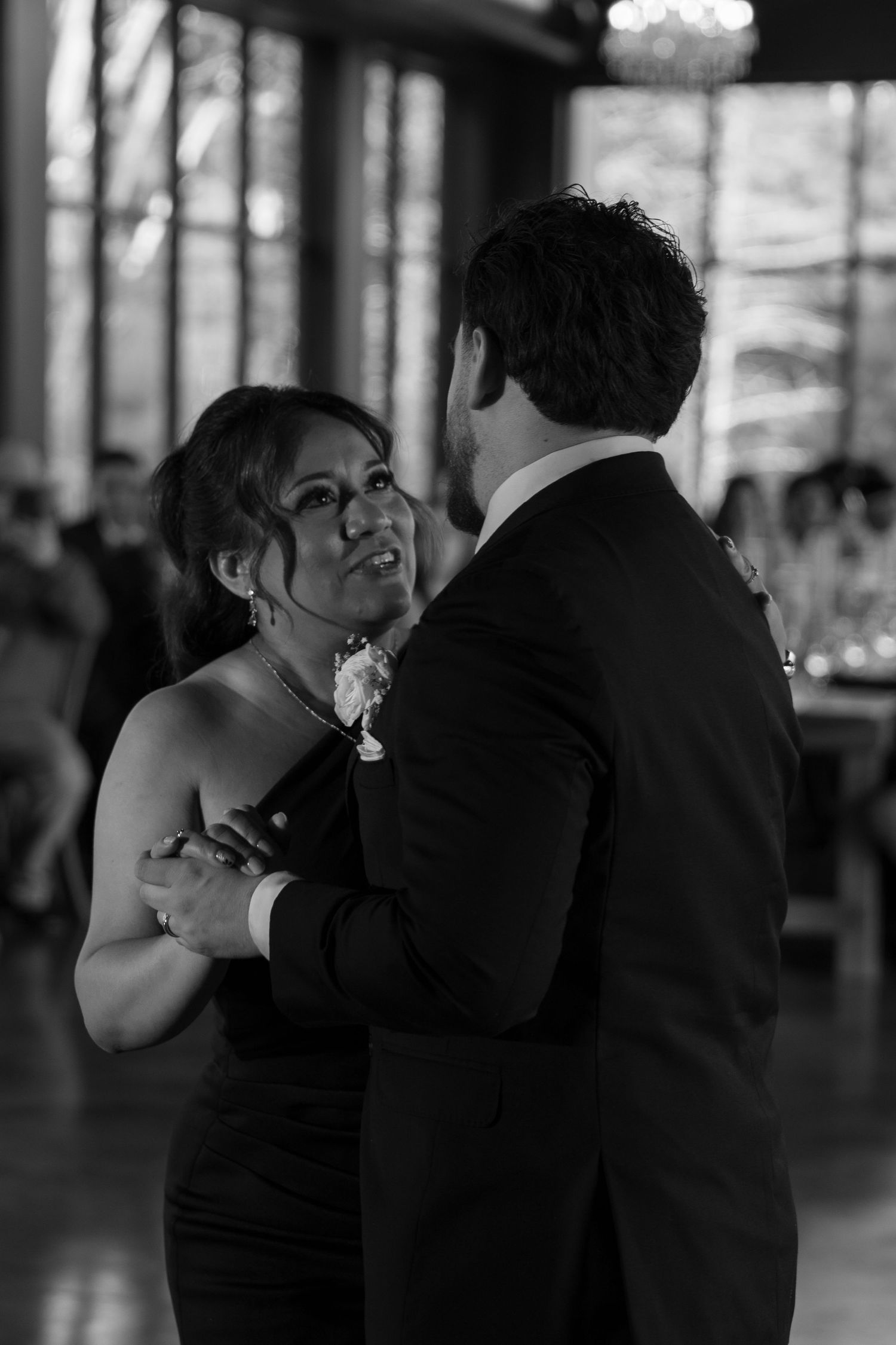 Groom shares tender mother-son dance with emotional mother in black and white wedding reception photograph.