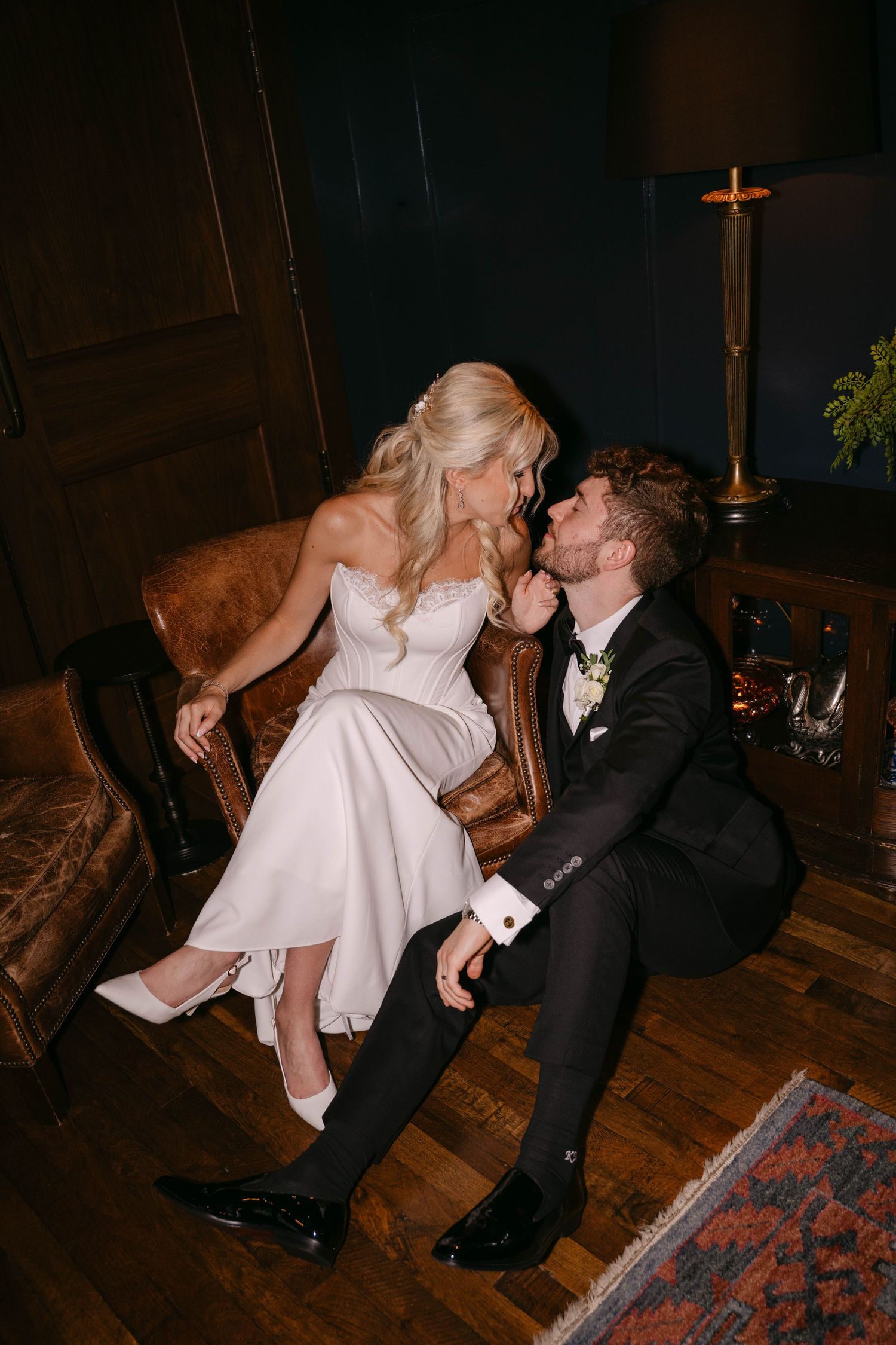 Bride and groom sharing intimate moment on leather sofa in dark elegant room.
