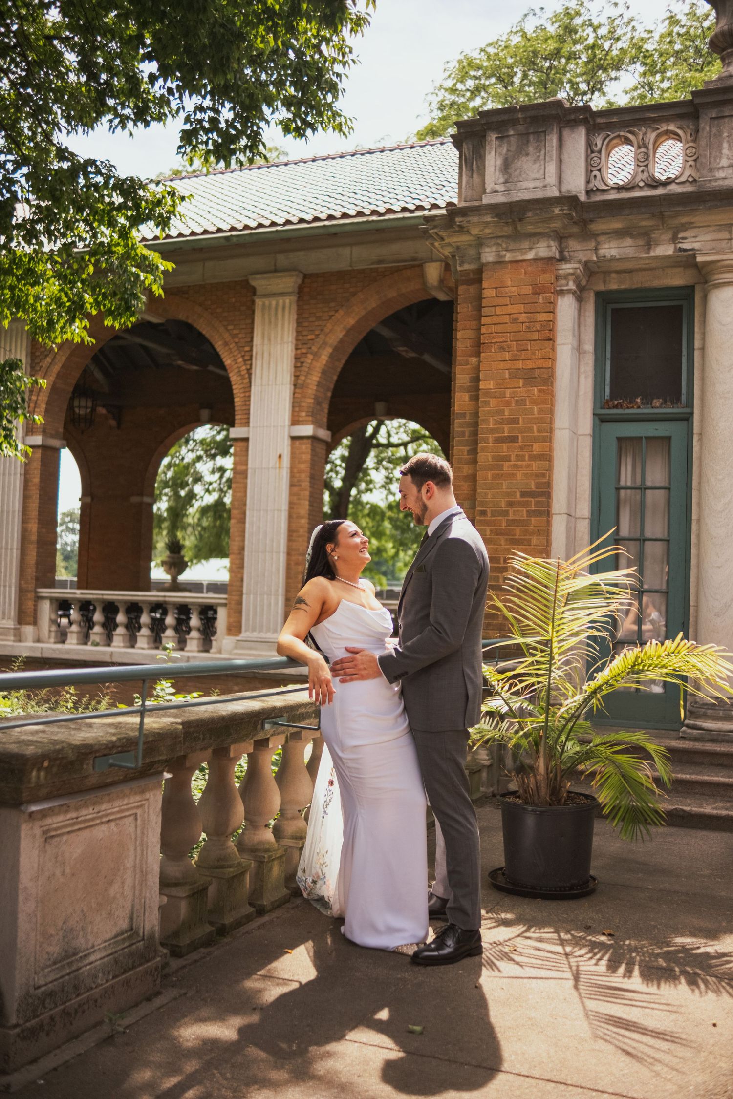 Bride and groom embracing on sunlit terrace of historic brick building with arched columns.