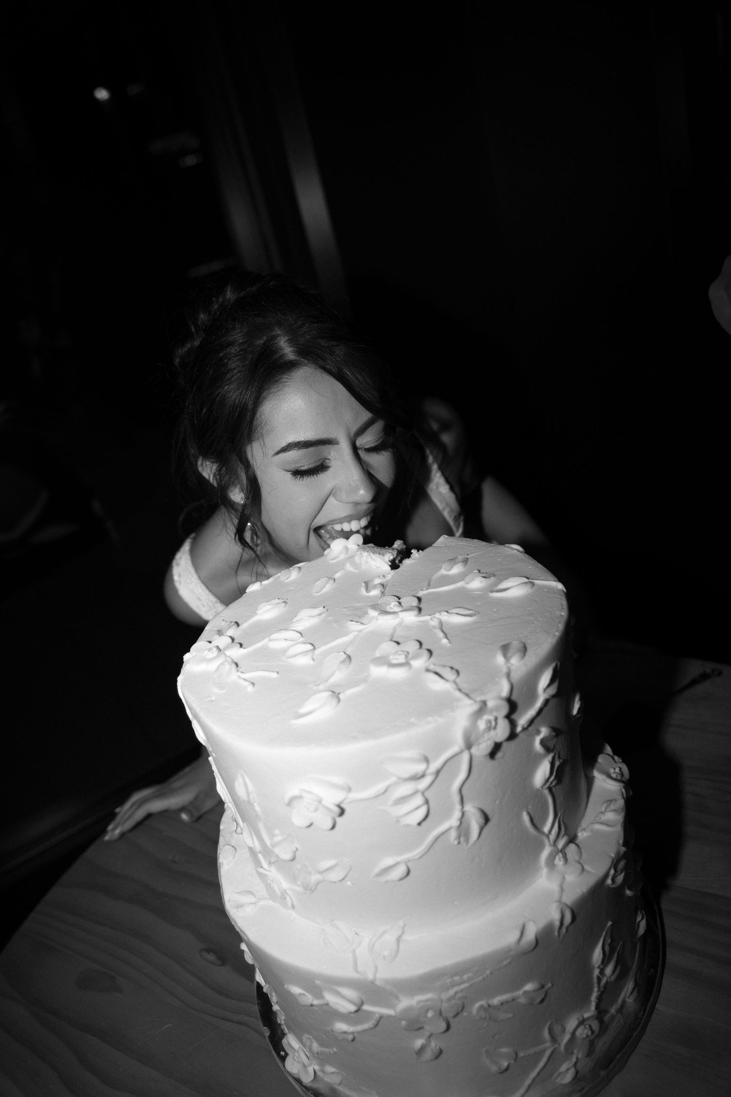 Black and white photo of a bride joyfully biting into her elegant two-tier white floral wedding cake.