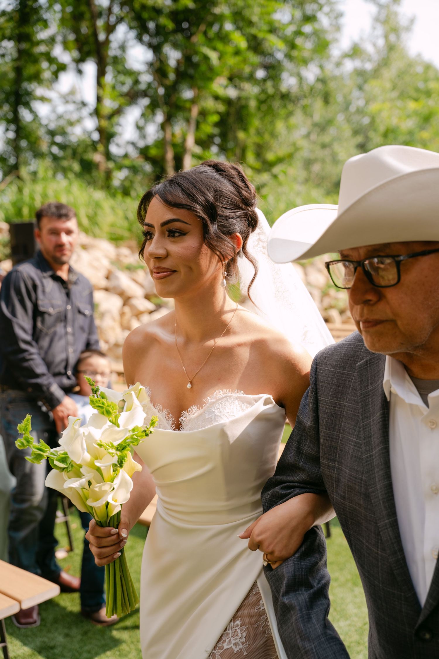 Beautiful bride in strapless wedding dress holding white calla lilies walks with father in cowboy hat.