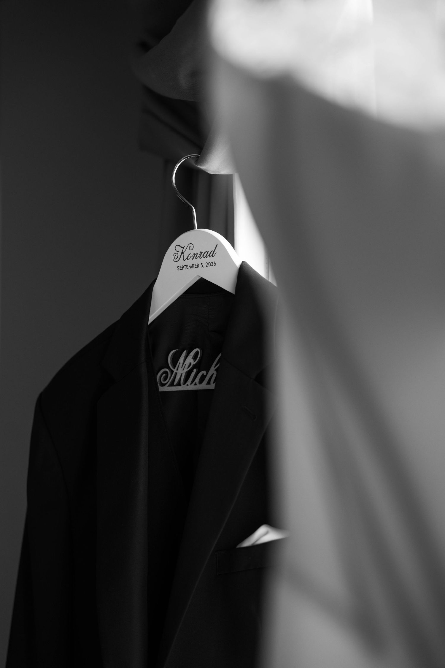 Black and white photo of a dark suit jacket on a personalized wedding hanger engraved with the name Conrad.