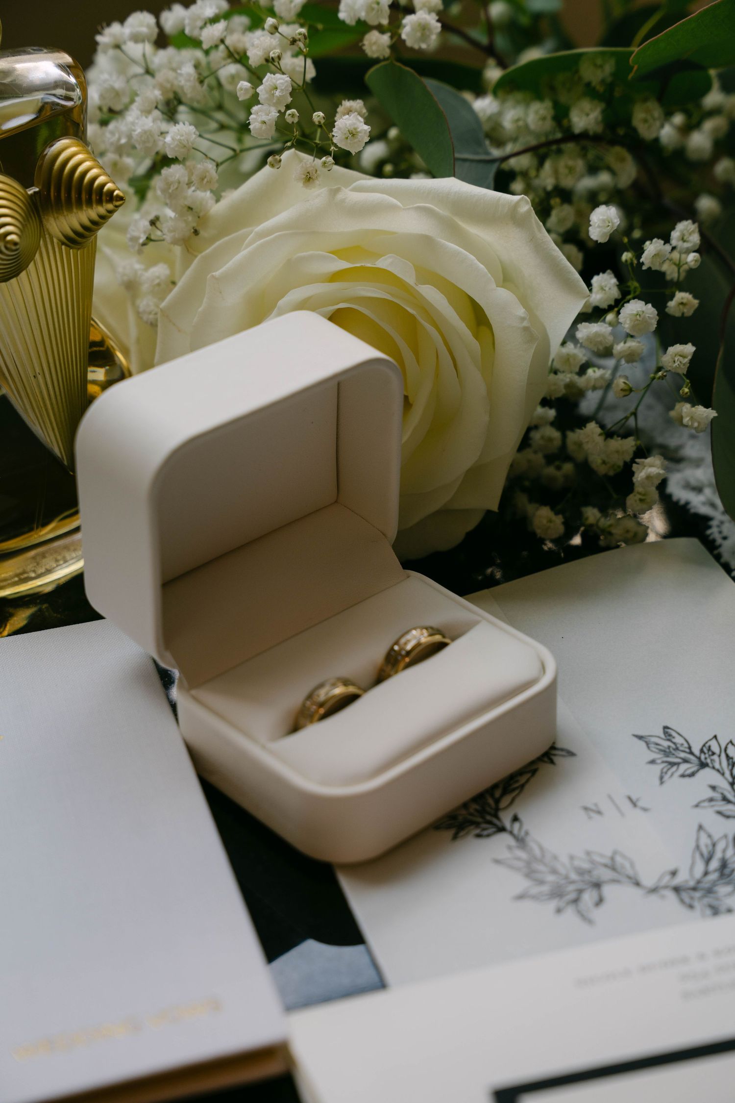 Two gold wedding bands displayed in a white velvet ring box beside a white rose bouquet and wedding invitation.