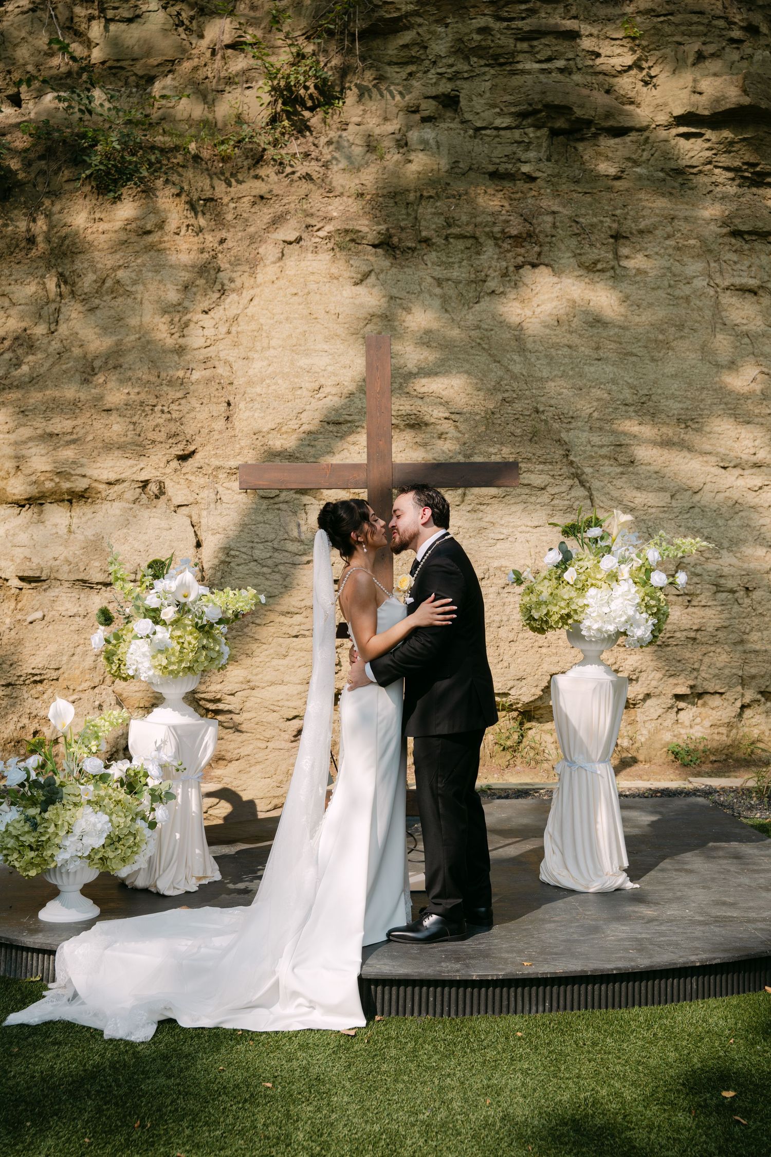 Bride in white gown with veil and groom in black suit kiss before wooden cross at ceremony.