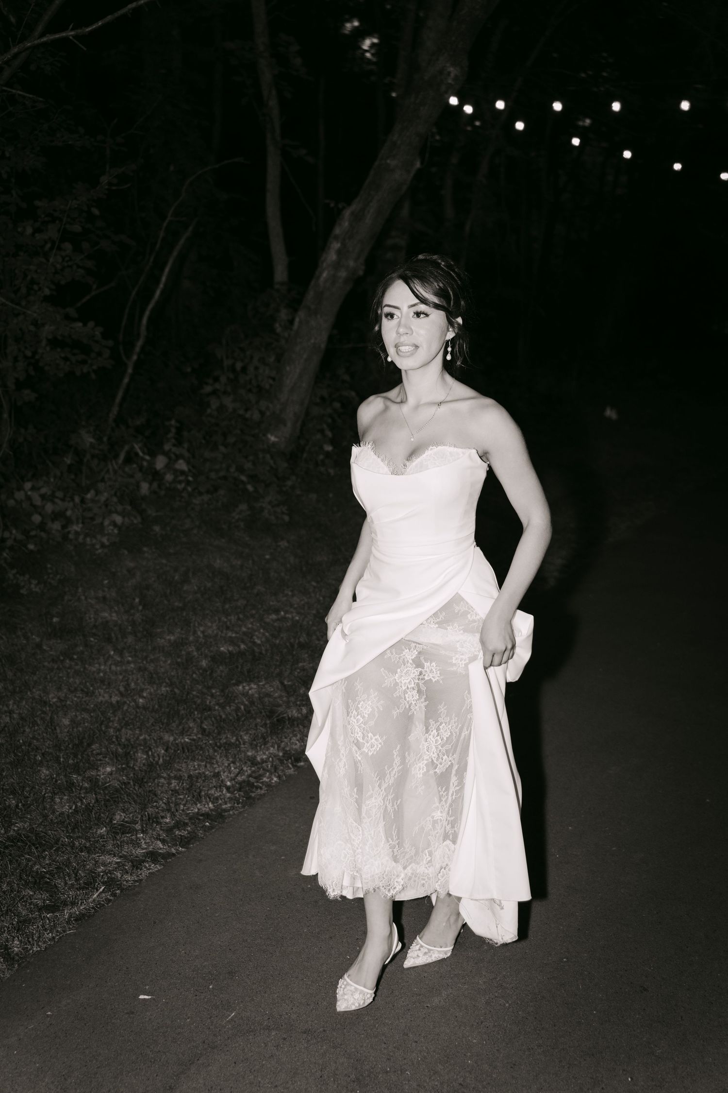 Black and white photo of smiling bride in strapless white lace gown on tree-lined path at night.