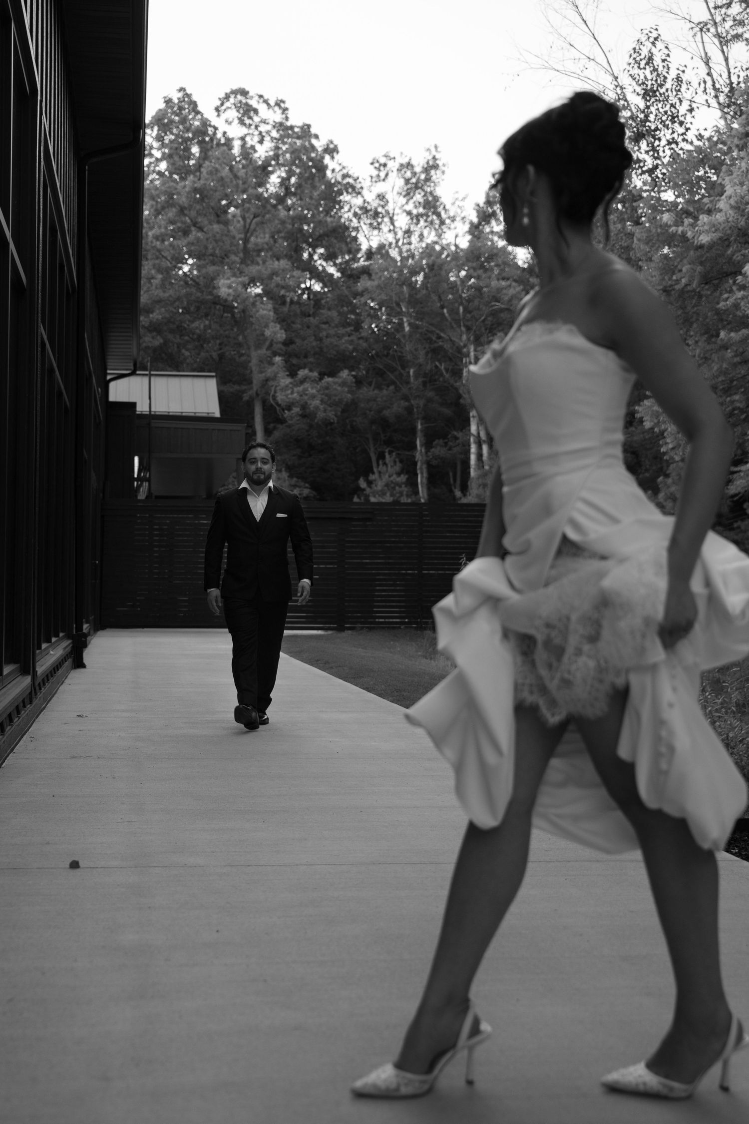 Bride in white strapless gown with lace detail walks toward groom in black suit on outdoor pathway.
