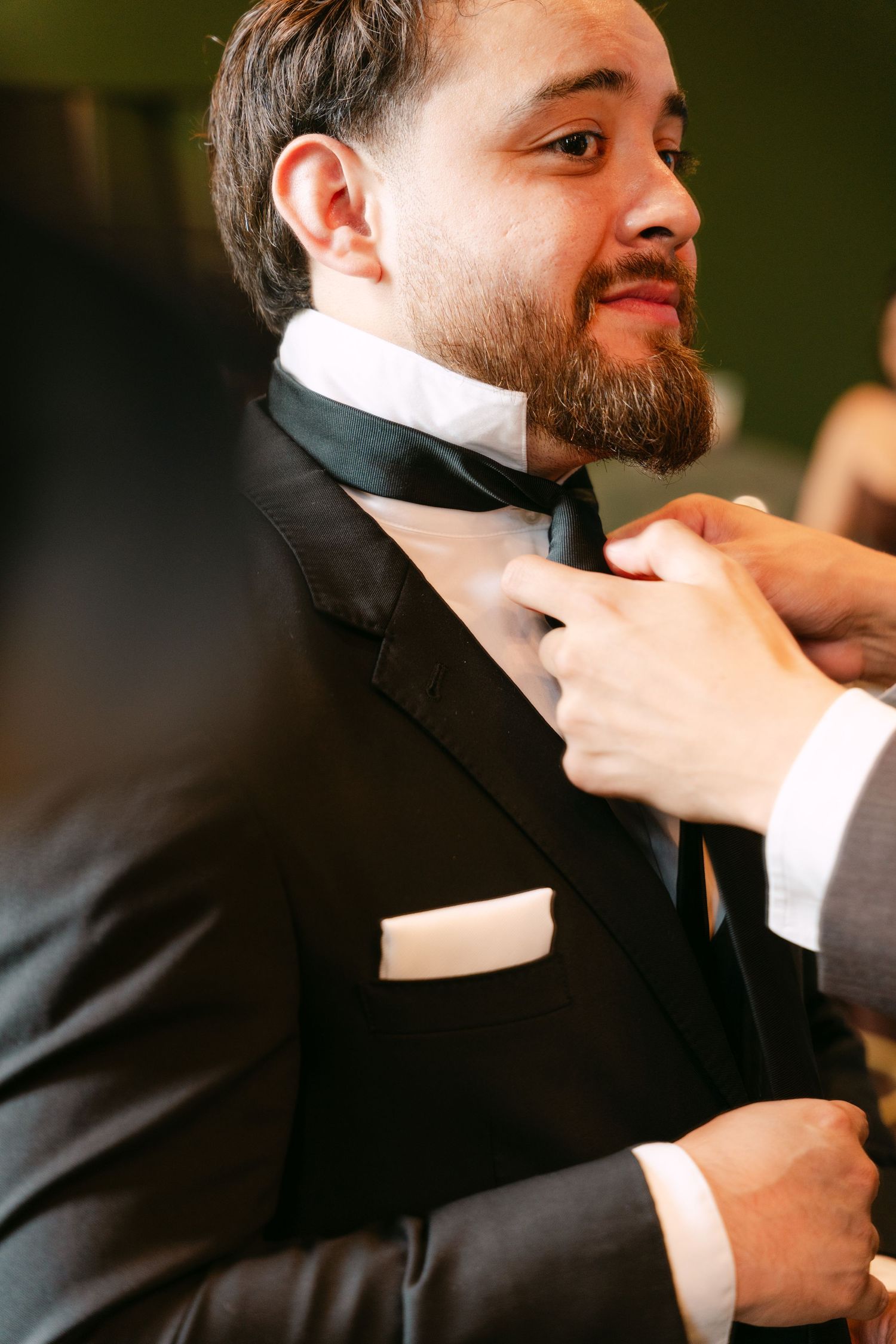 Groom in black tuxedo smiling as his tie is adjusted during wedding preparations.