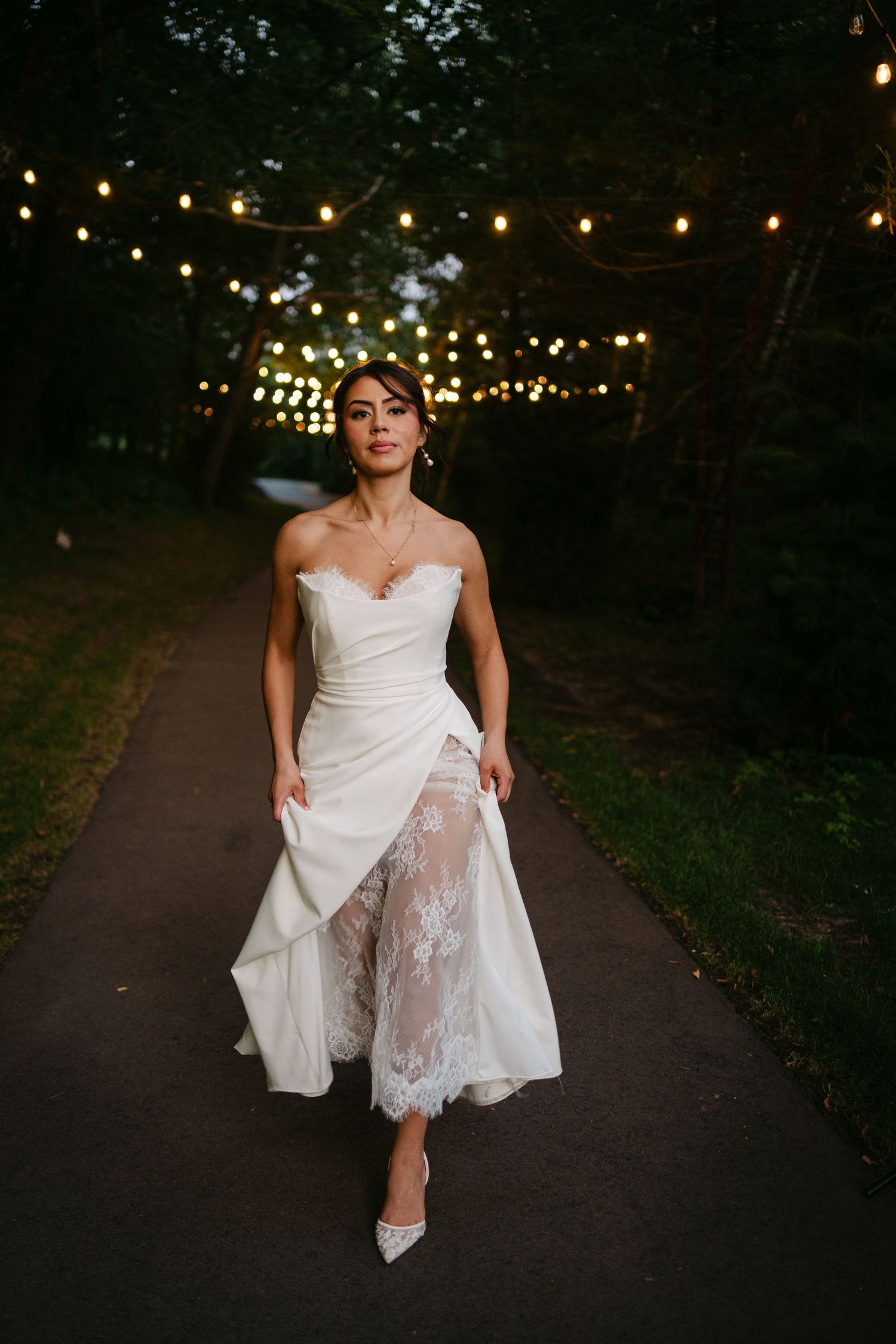 Elegant bride in strapless white lace wedding dress strolls down tree-lined path under string lights.