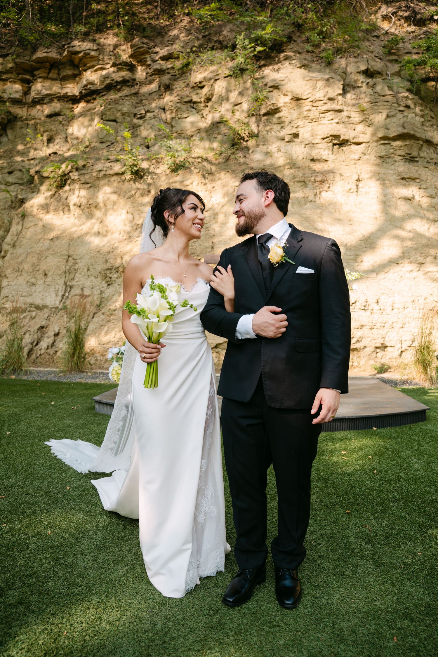 Happy bride holding white bouquet and groom in black suit laughing together outdoors by rocky cliff.