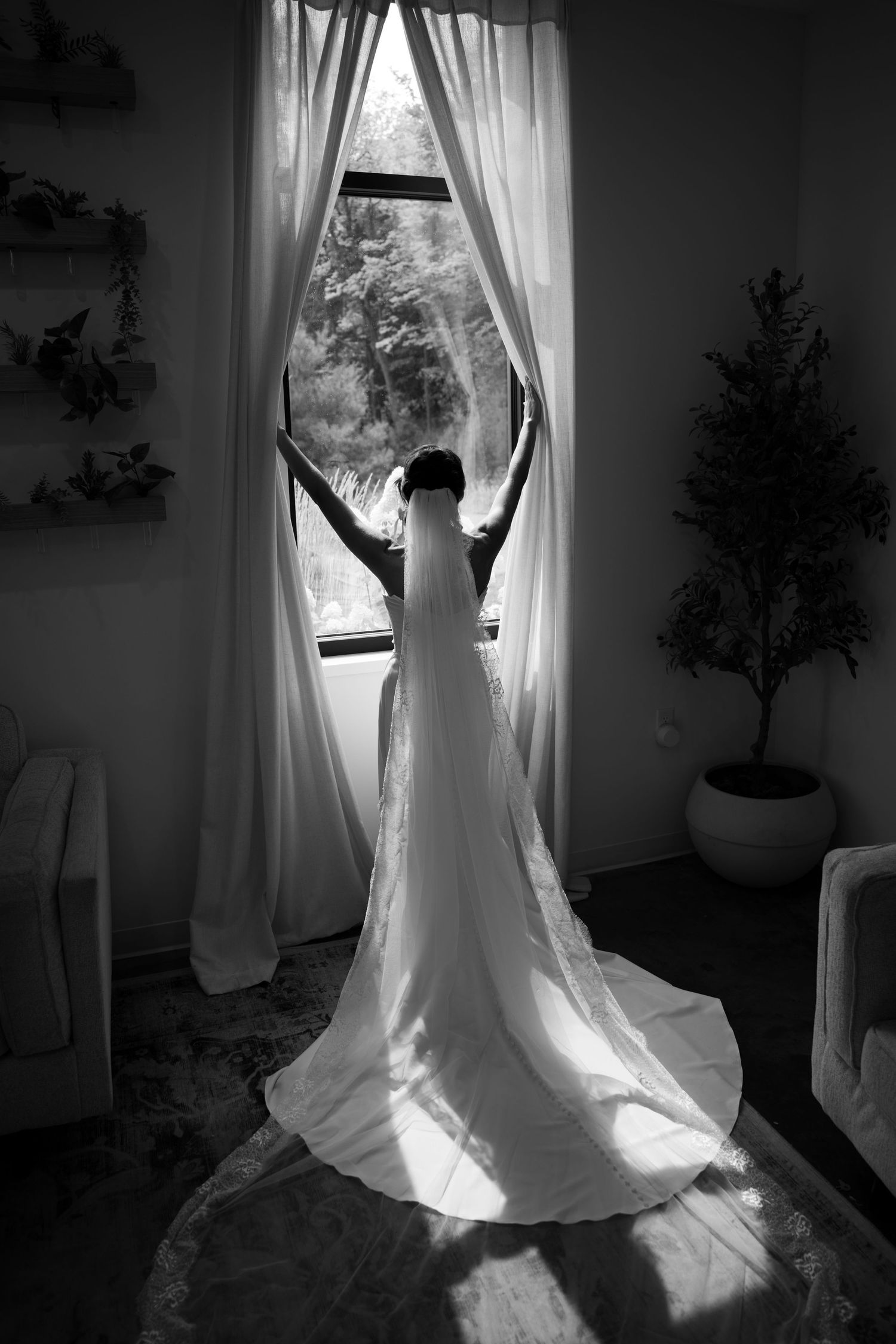 Black and white photo of bride in cathedral veil gown reaching toward curtains at a window.