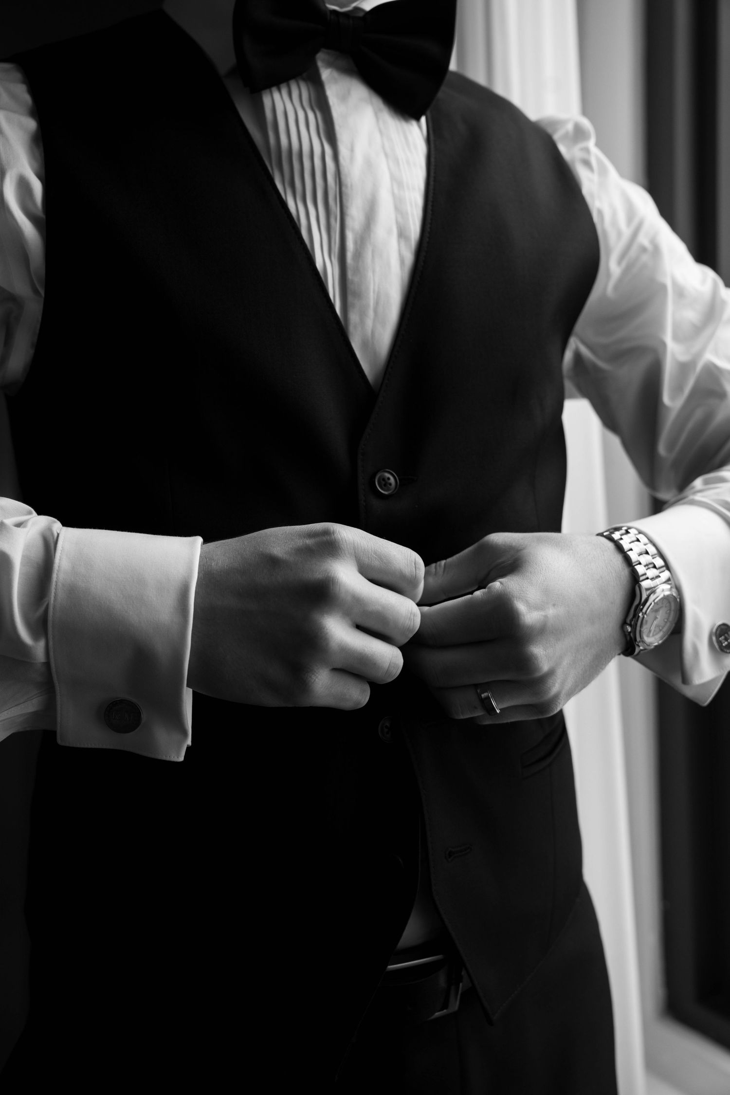 Groom in black vest and bow tie fastening cufflinks, wearing a watch, in elegant black and white wedding photo.