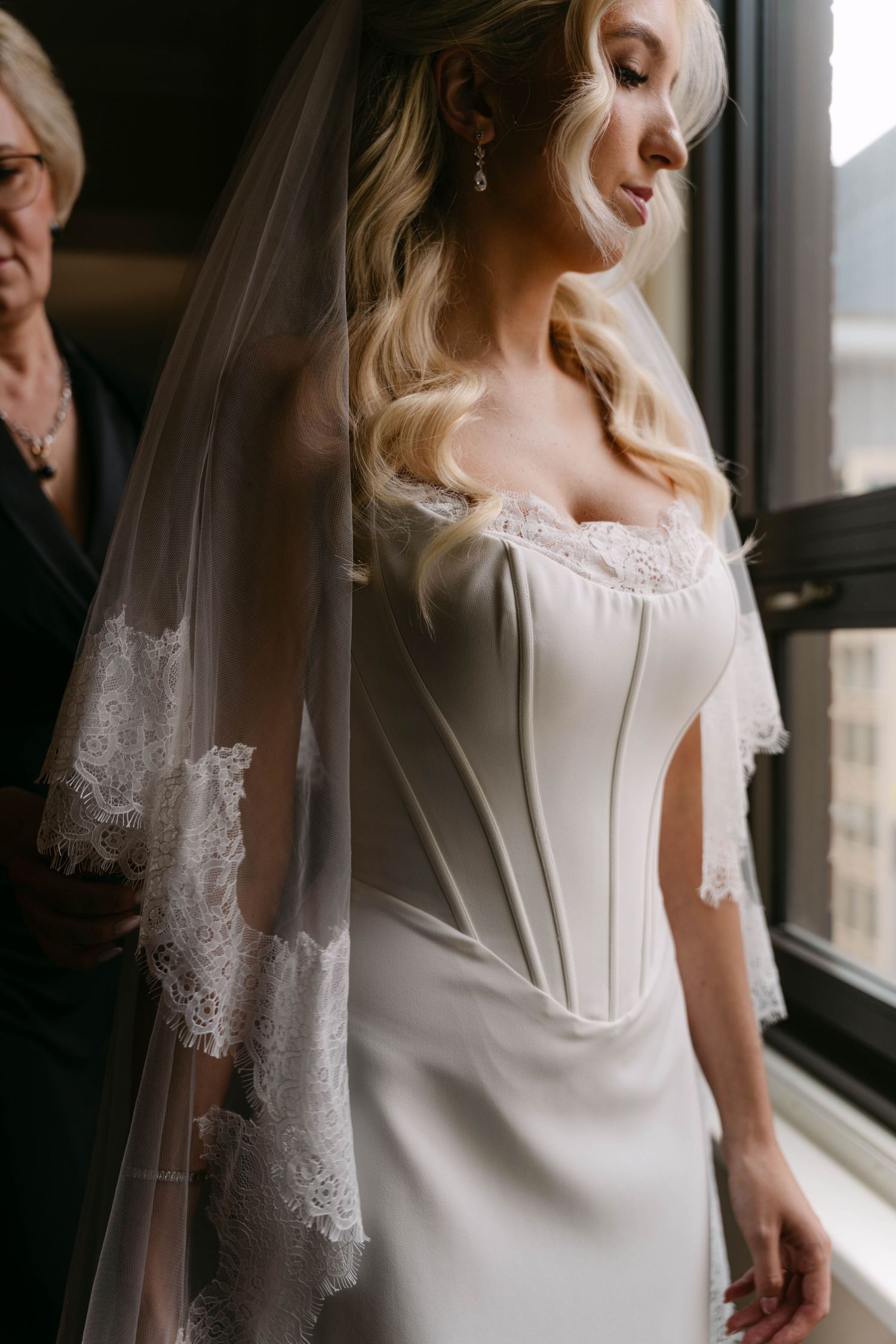 Blonde bride in corset wedding dress and lace veil gazes down by window while being helped by older woman.