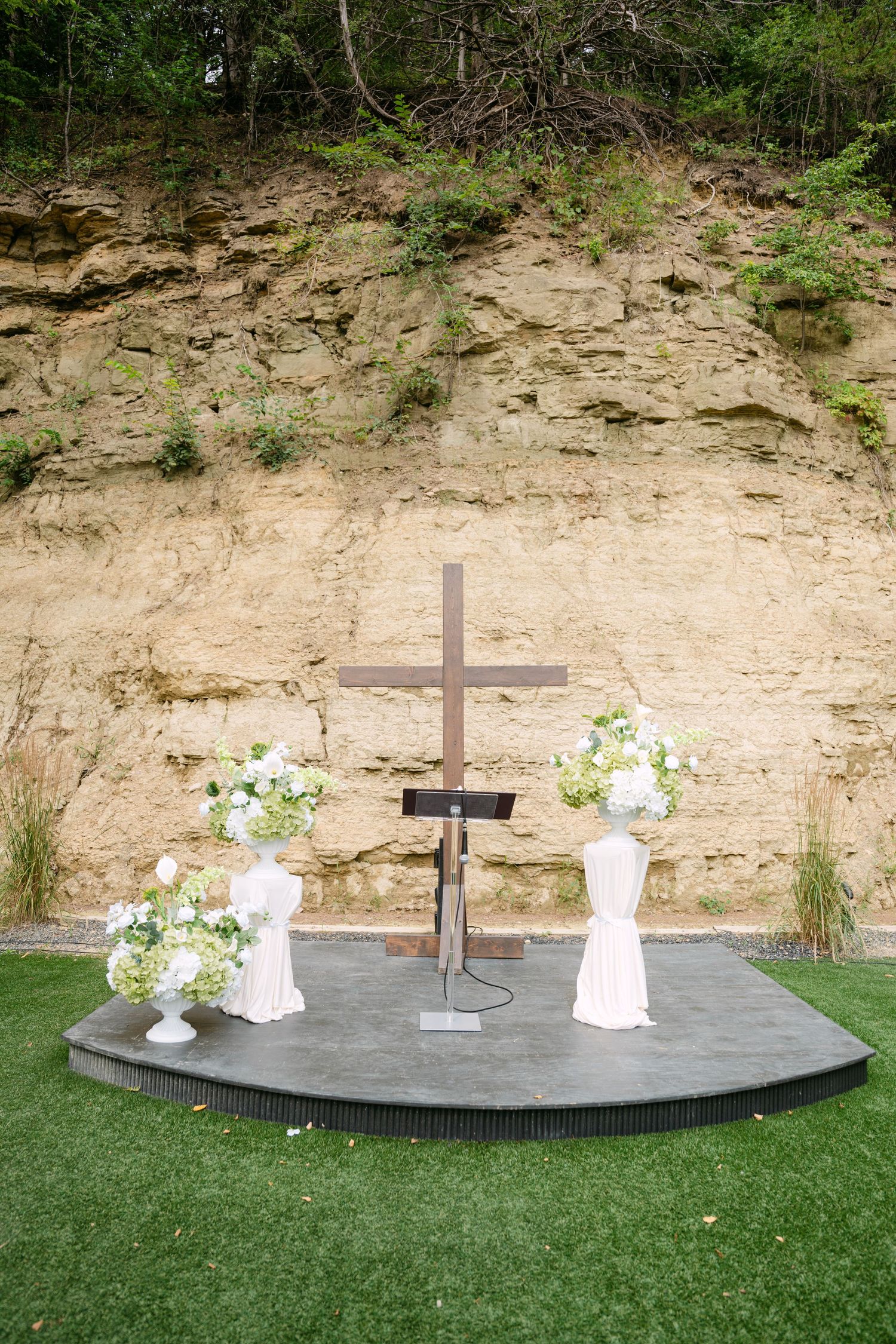 Outdoor wedding altar with wooden cross flanked by white floral arrangements against sandy rock wall.