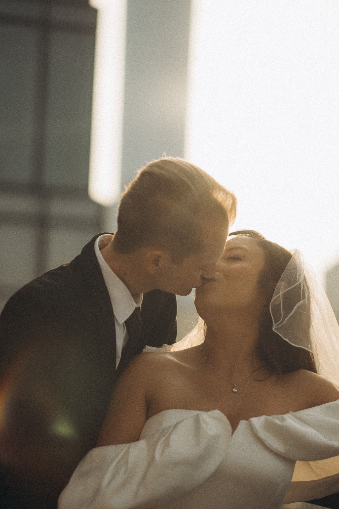 A bride and groom share a romantic kiss backlit by golden sunlight, with the bride wearing a white off-shoulder gown and veil.