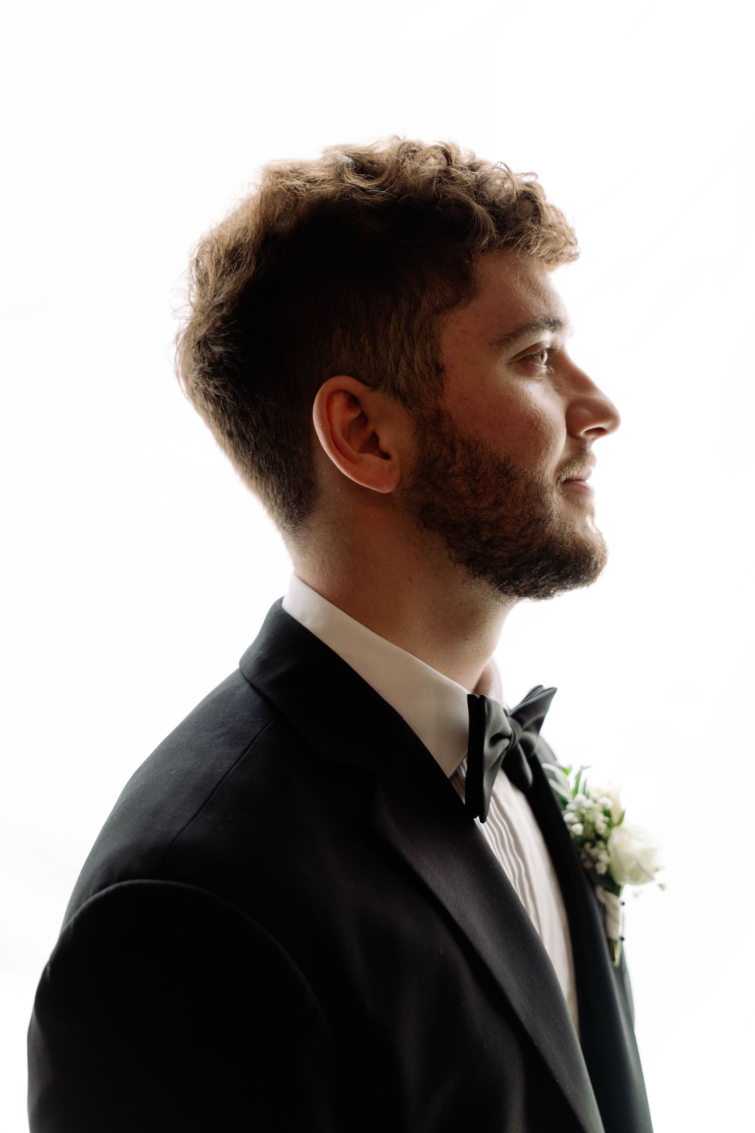 Groom in black tuxedo with boutonniere smiling in profile against white background.
