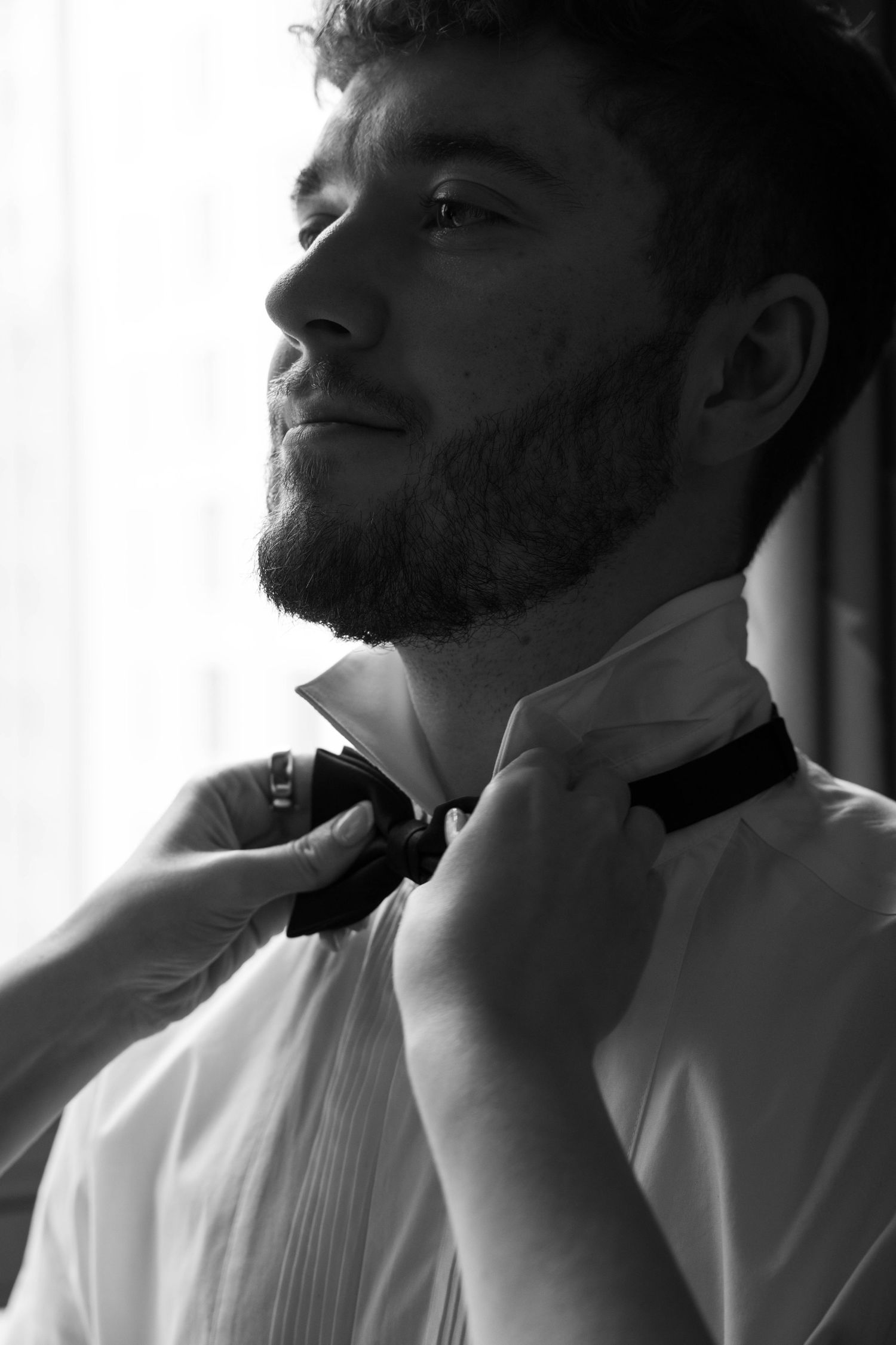 Groom smiles as hands adjust his bow tie in a dramatic black and white close-up wedding photo.