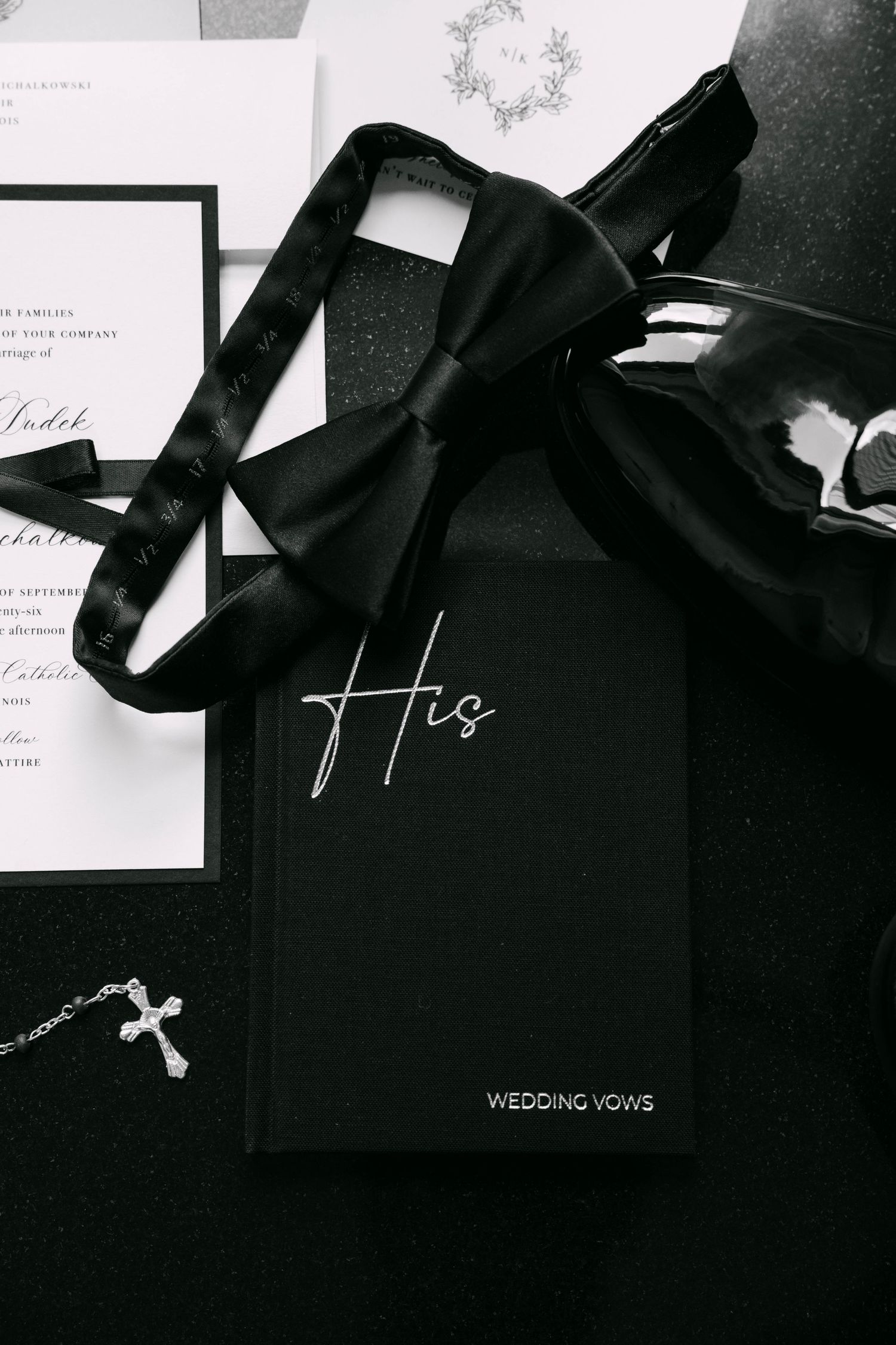 Black wedding vow book labeled His with bow tie and cross necklace on dark background.
